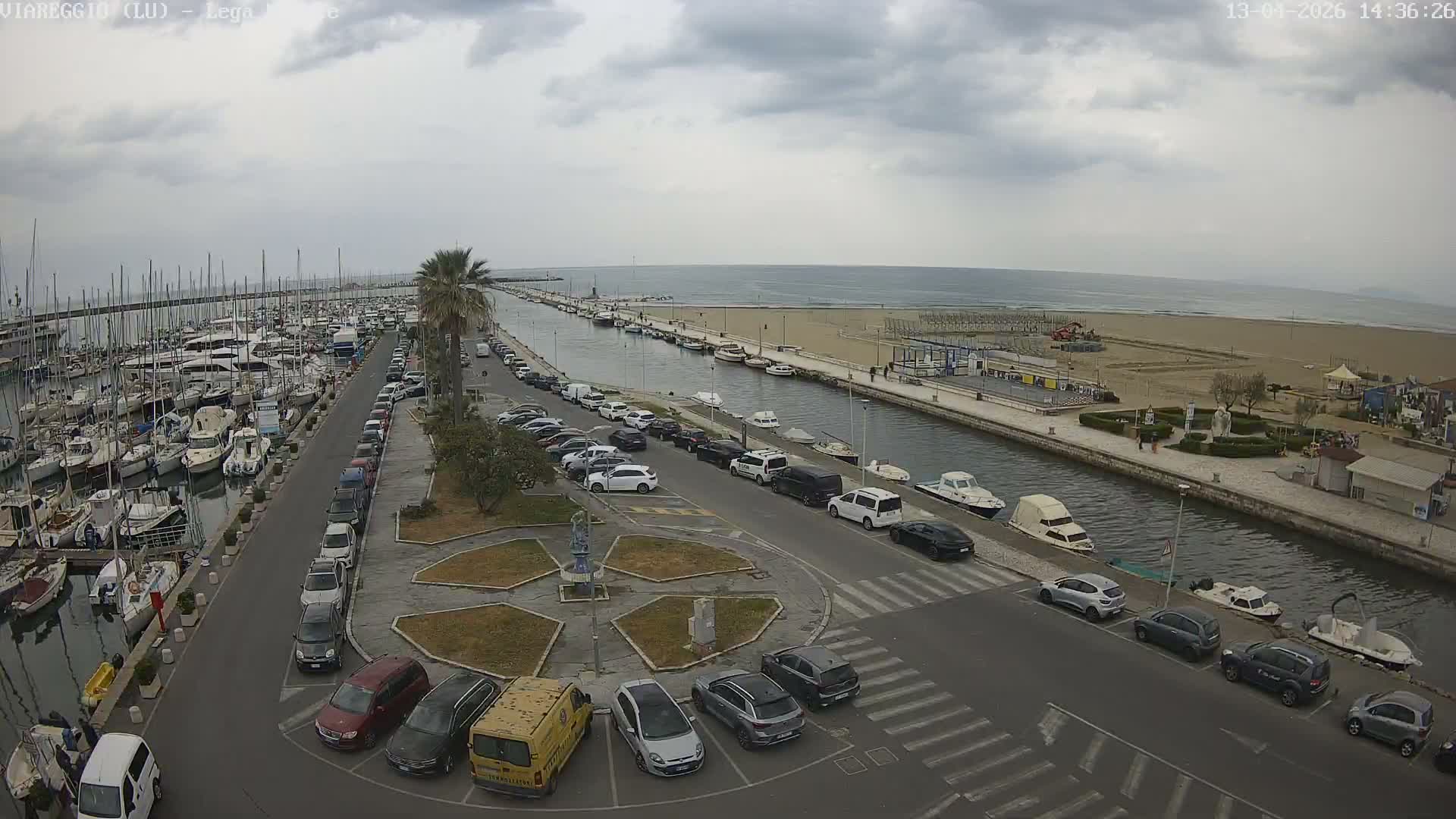 Under a cloudy sky, a wide panorama captures a bustling marina filled with numerous sailboats and yachts on the left, a parallel canal with smaller boats, a road, a sandy beach, and the calm sea stretching to the horizon on the right.