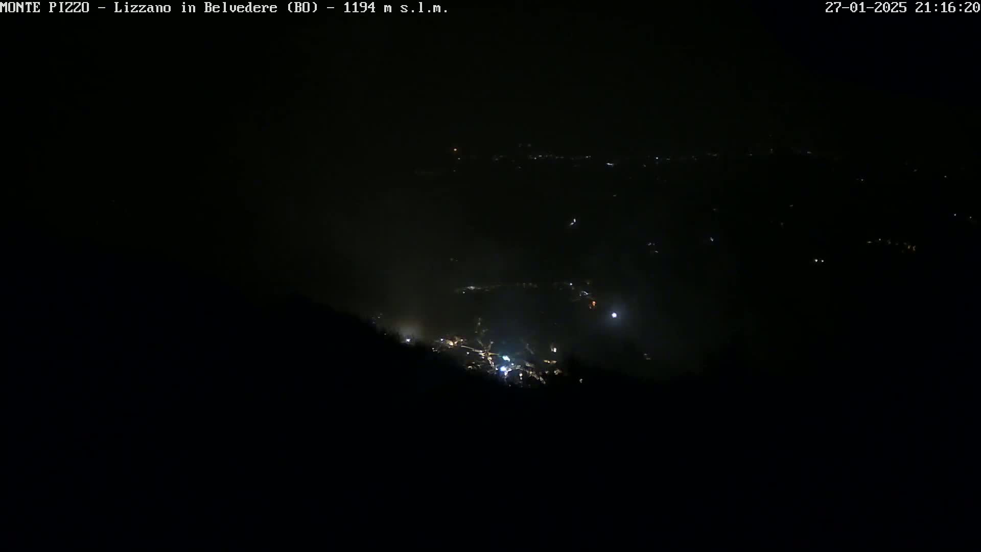 A nighttime, high-angle view of a dark valley with scattered lights below, possibly a town or village, under a clear, dark sky.