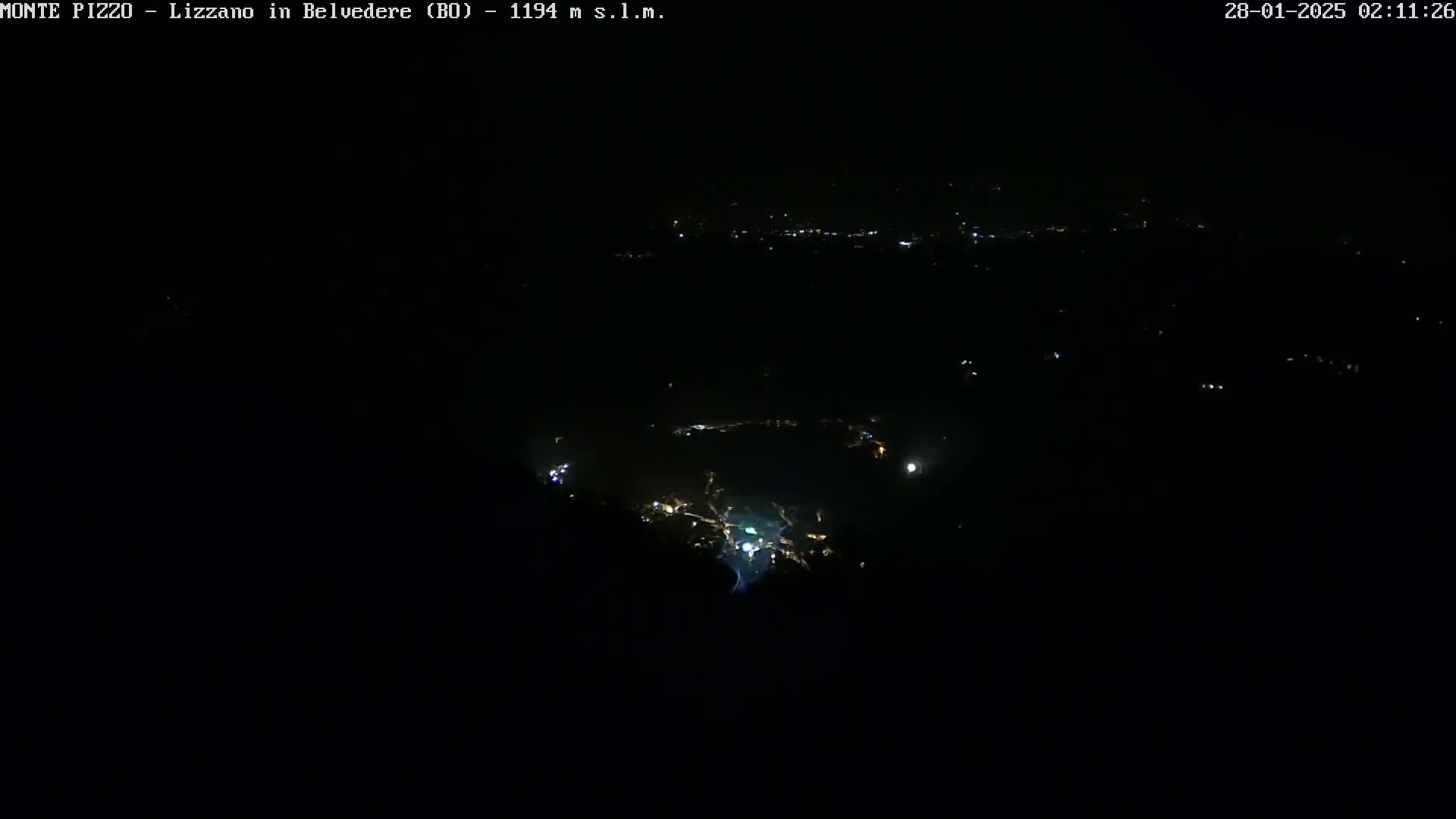 A nighttime, high-angle view shows a dark landscape with scattered lights from a town below.