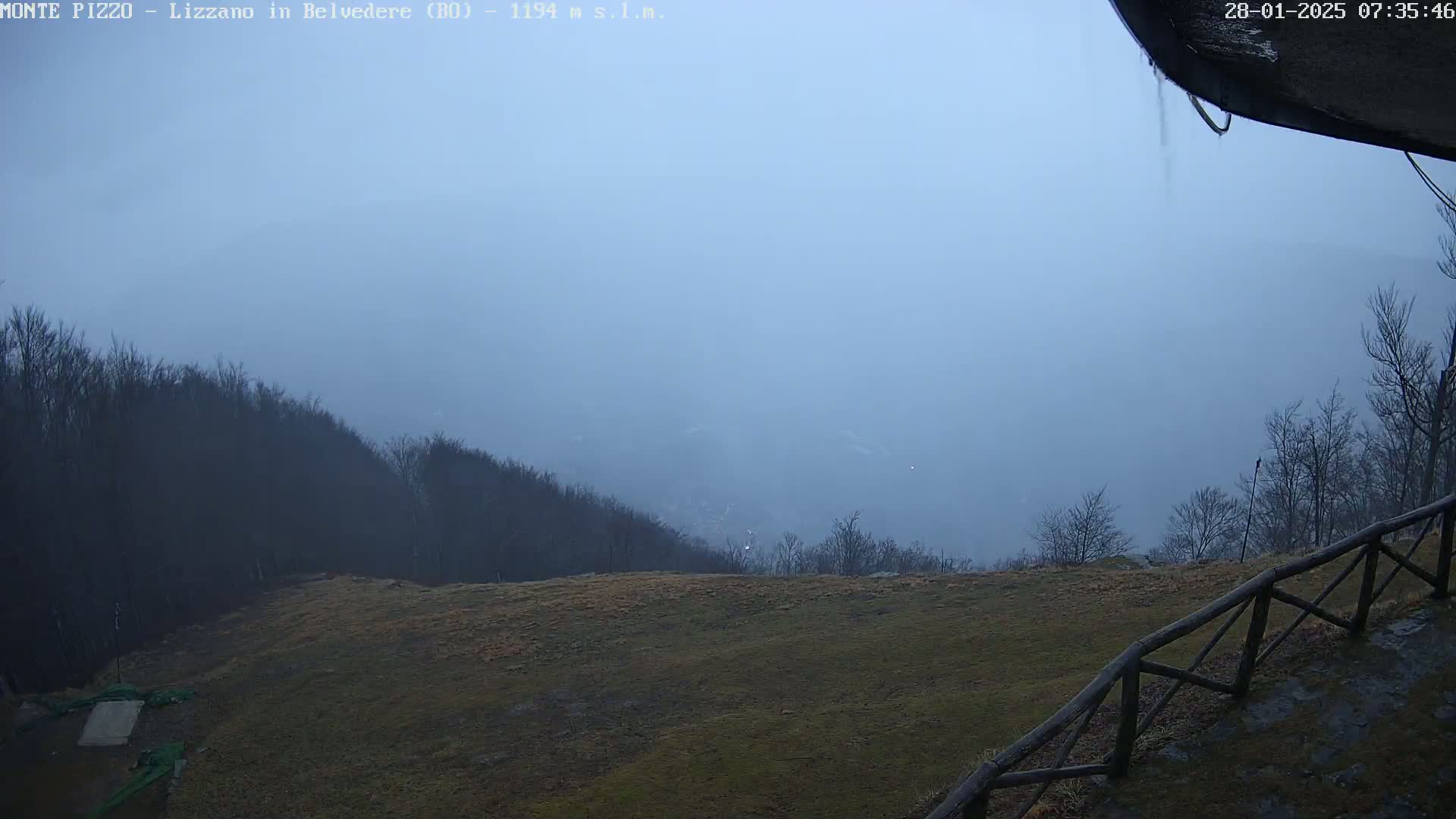 A foggy, overcast view from a hilltop shows a grassy area with a wooden fence in the foreground and a distant, hazy valley surrounded by bare trees.