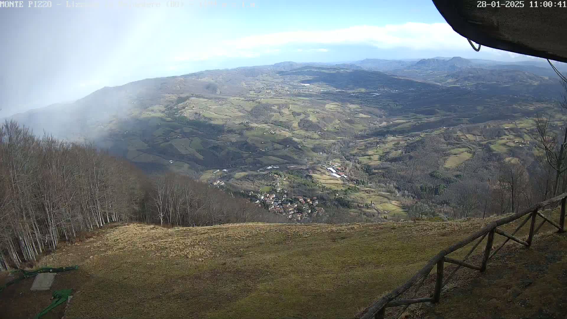 A panoramic view from a mountaintop shows a valley with patchy fields and a small village below, partially obscured by fog, under a partly cloudy sky.