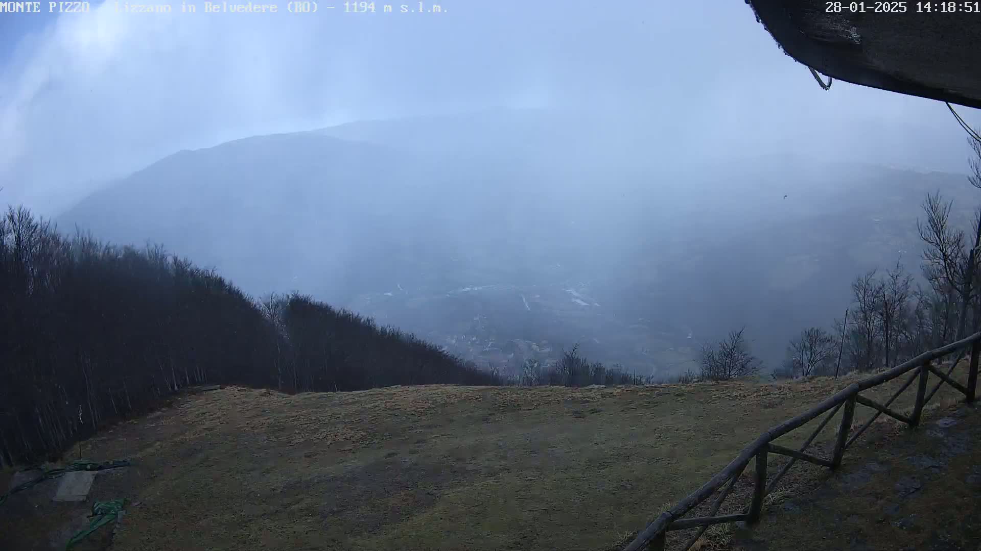 A hazy, overcast view from a mountaintop shows a valley shrouded in mist, with bare trees lining the foreground and a wooden fence along the edge of a grassy area.