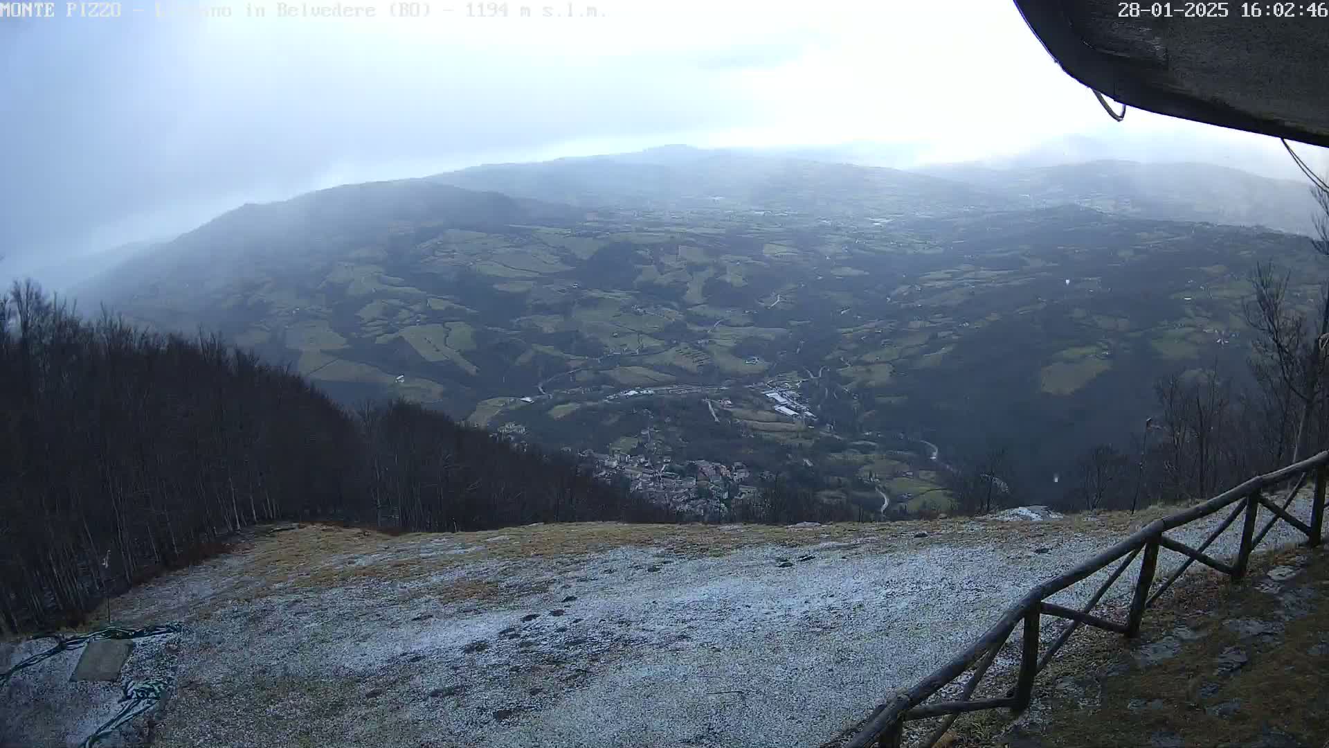 A snow-dusted hillside overlooks a vast valley of farmland and a small town under an overcast sky.