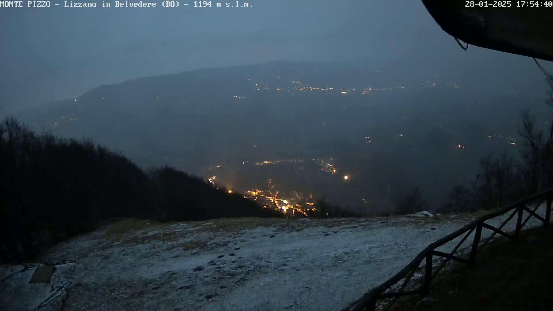 A snow-dusted, dimly lit landscape at night shows a valley with scattered lights below a hilltop observation point.