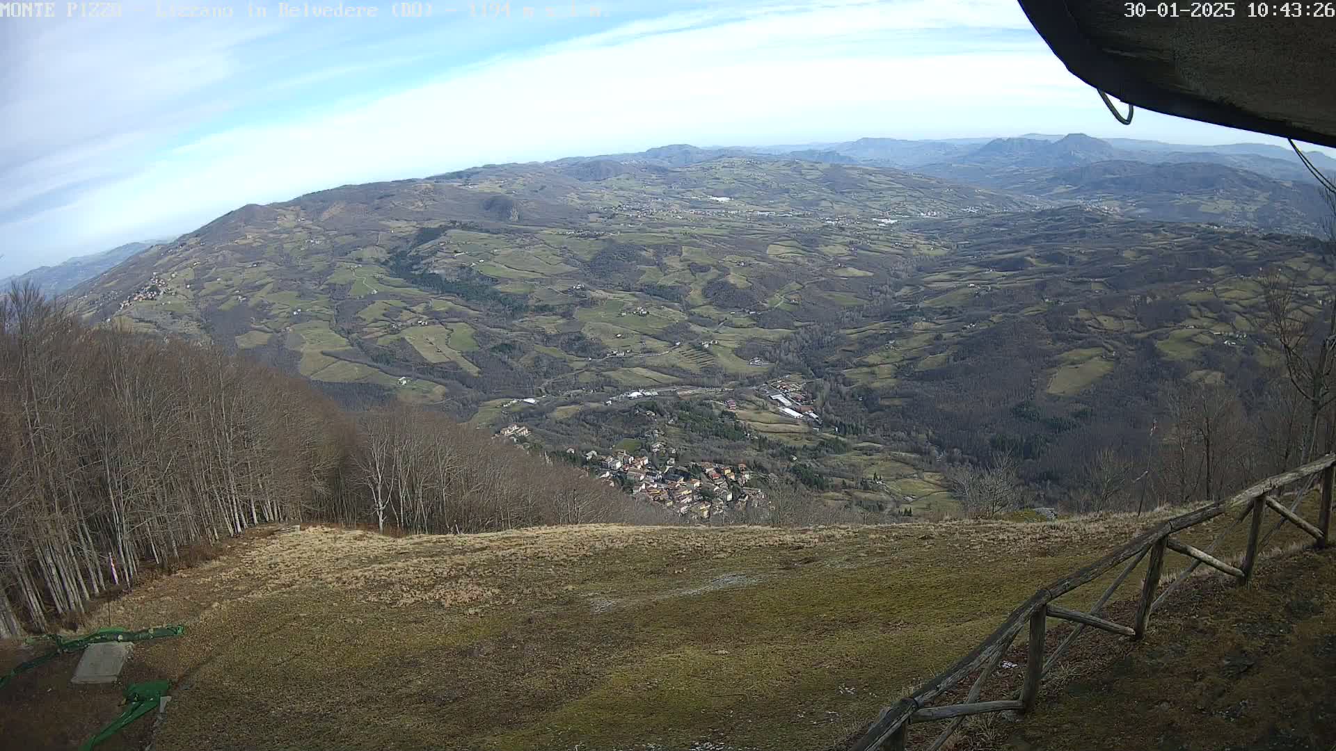 A high-angle view shows a vast, hilly landscape under a partly cloudy sky, with a small village nestled in a valley below, and a wooden fence in the foreground.