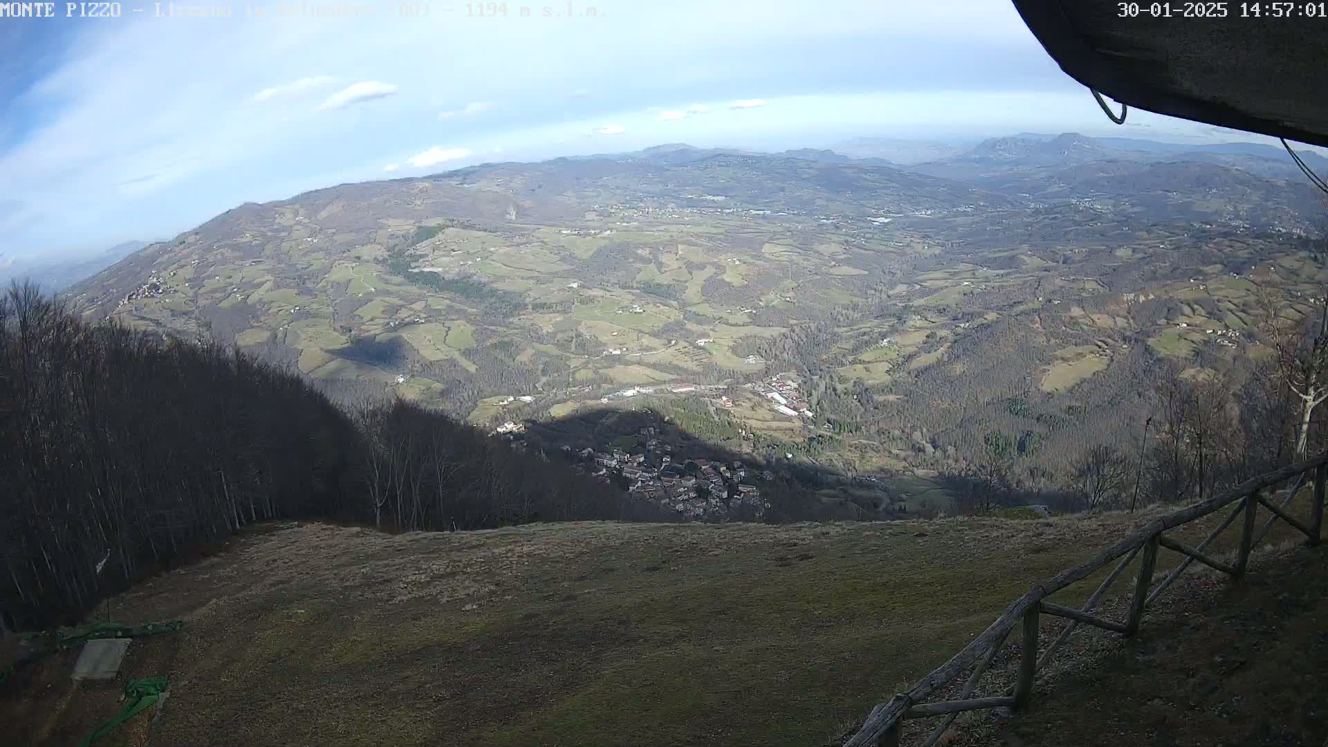 A panoramic view from a mountaintop shows a sprawling valley with farmland, forests, and a small village below, under a mostly sunny sky with a few clouds.