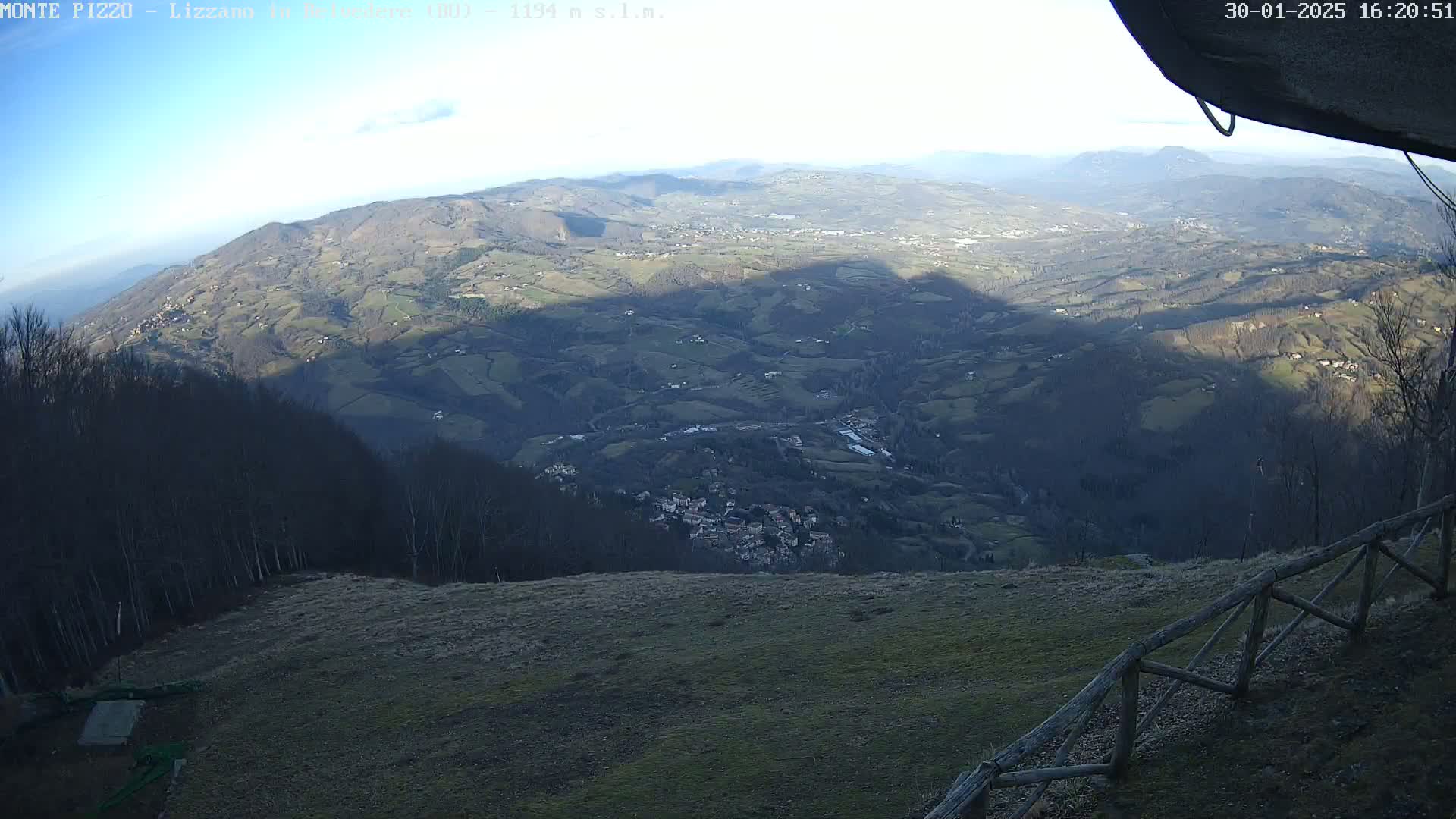 A high-angle, long shot shows a hilly landscape under a clear, bright sky, with a small village nestled in a valley below and a wooden fence in the foreground.