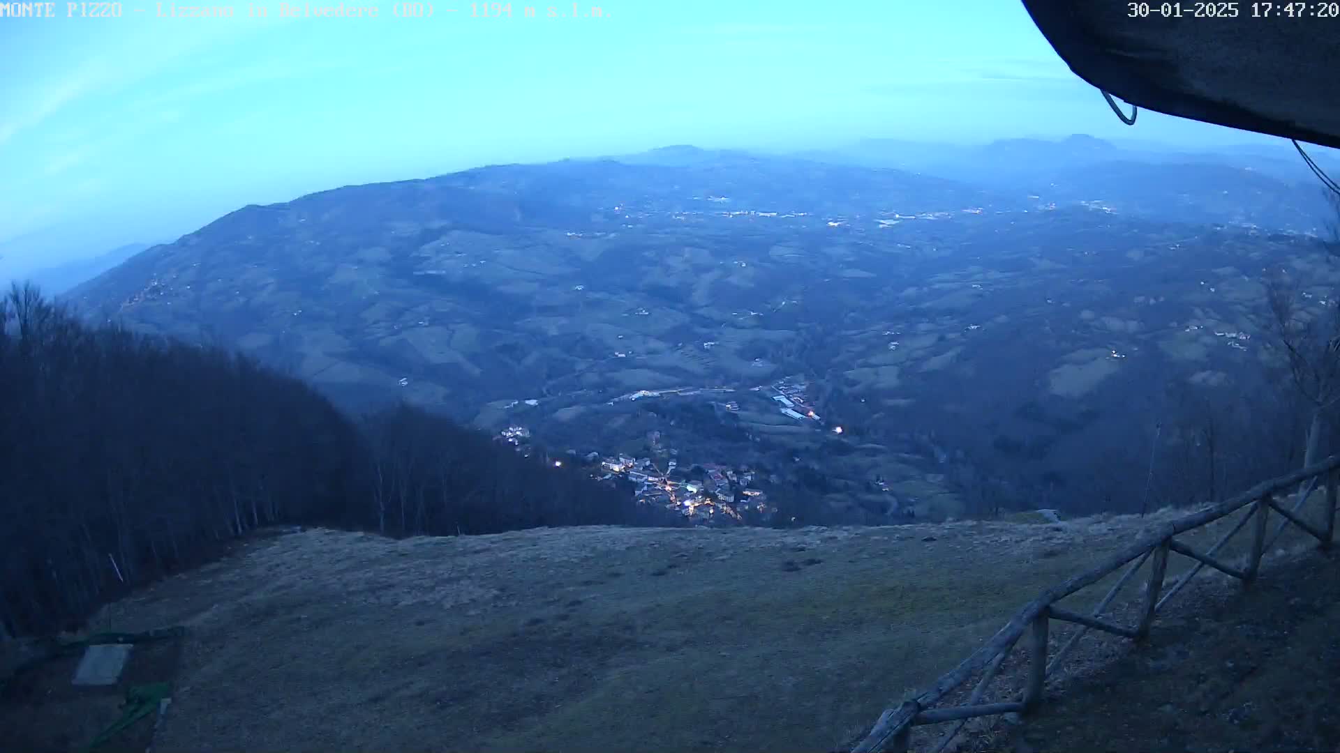 A high-angle, twilight view shows a valley town nestled among rolling hills and dark forests under a clear, dusky blue sky.