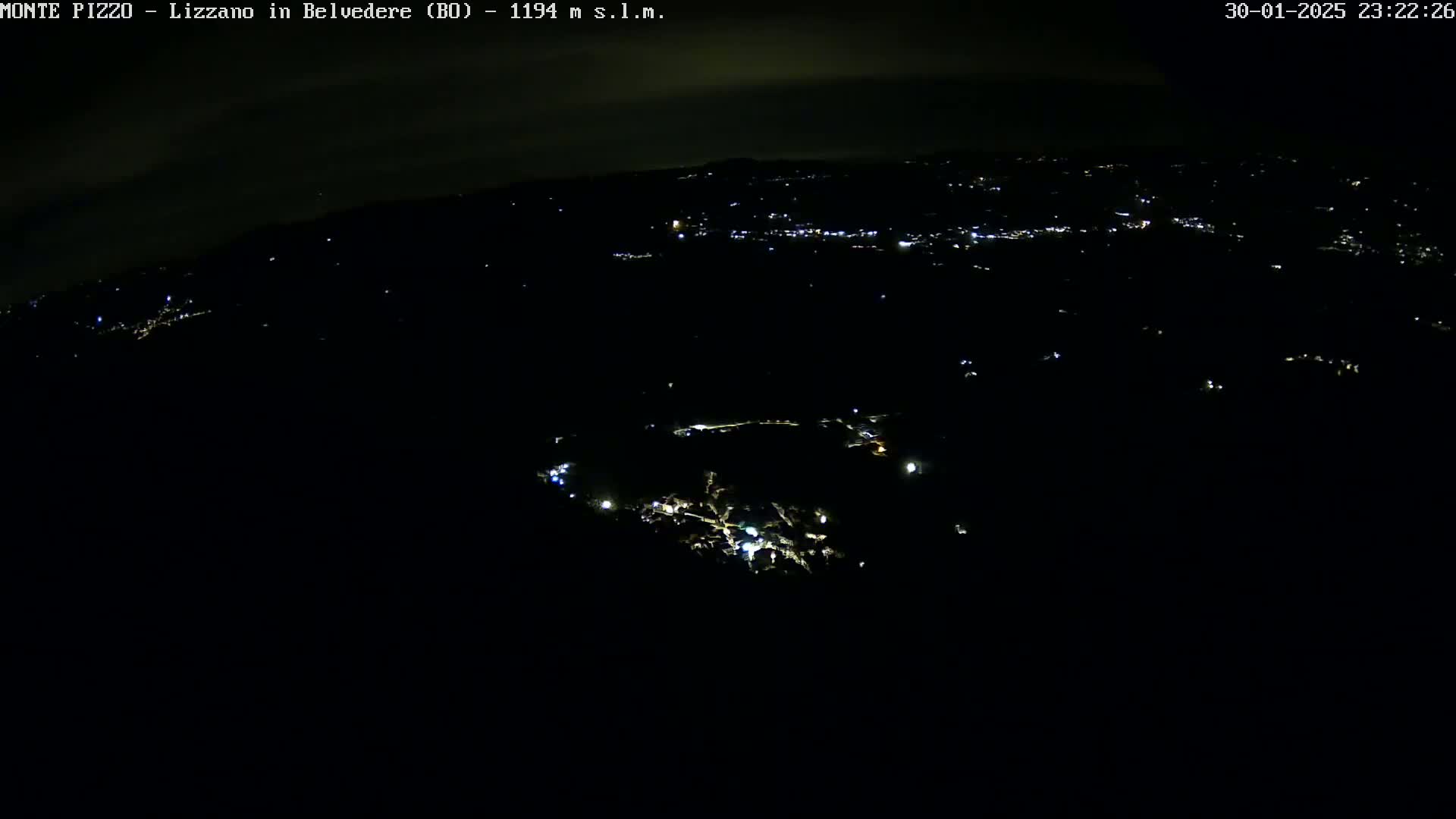 A nighttime aerial view shows a dark landscape with scattered lights from a town or village below, under a mostly clear night sky.