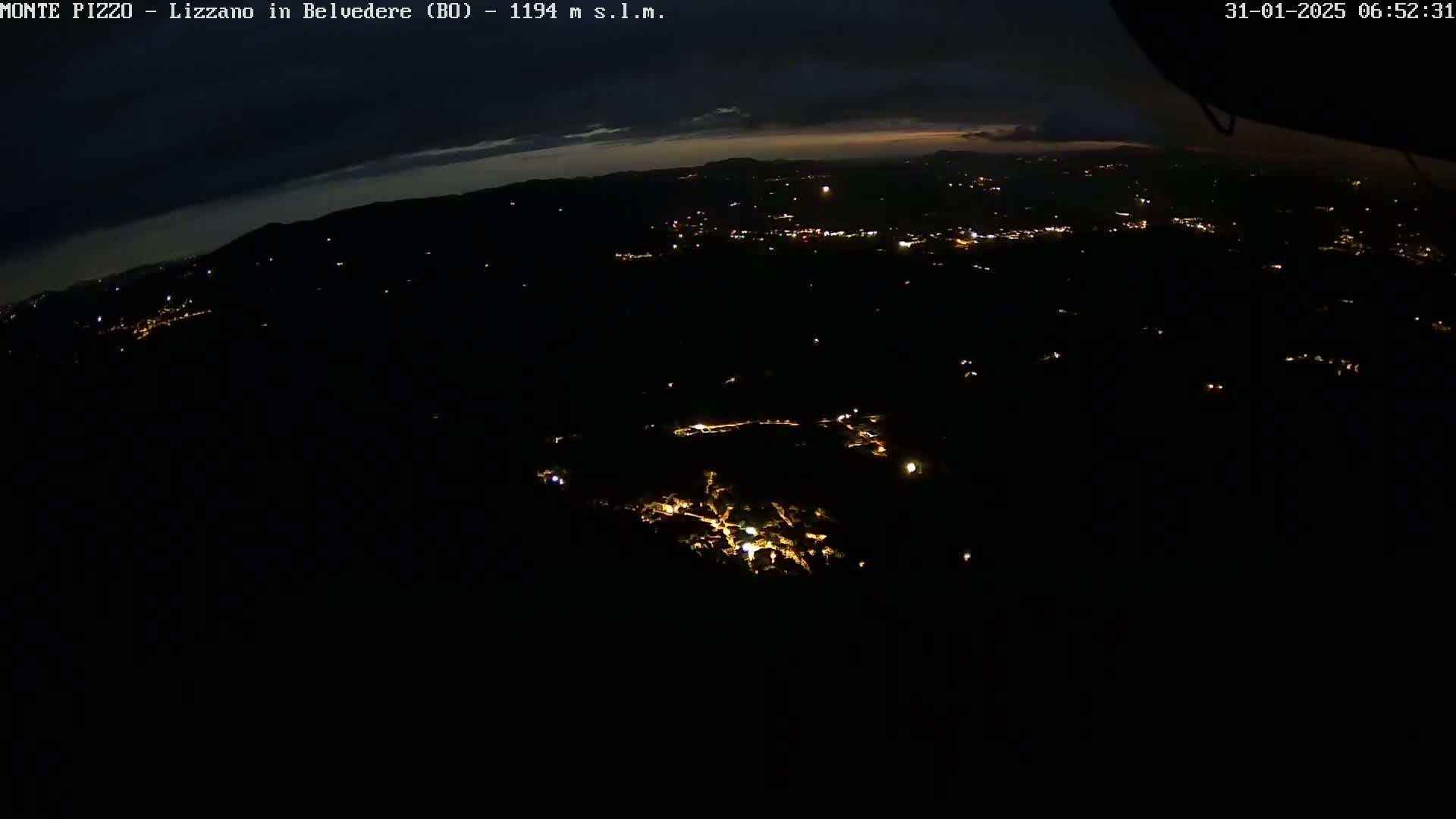 A nighttime, high-angle view shows a dark landscape with scattered lights from a town below, under a mostly clear, dark sky.