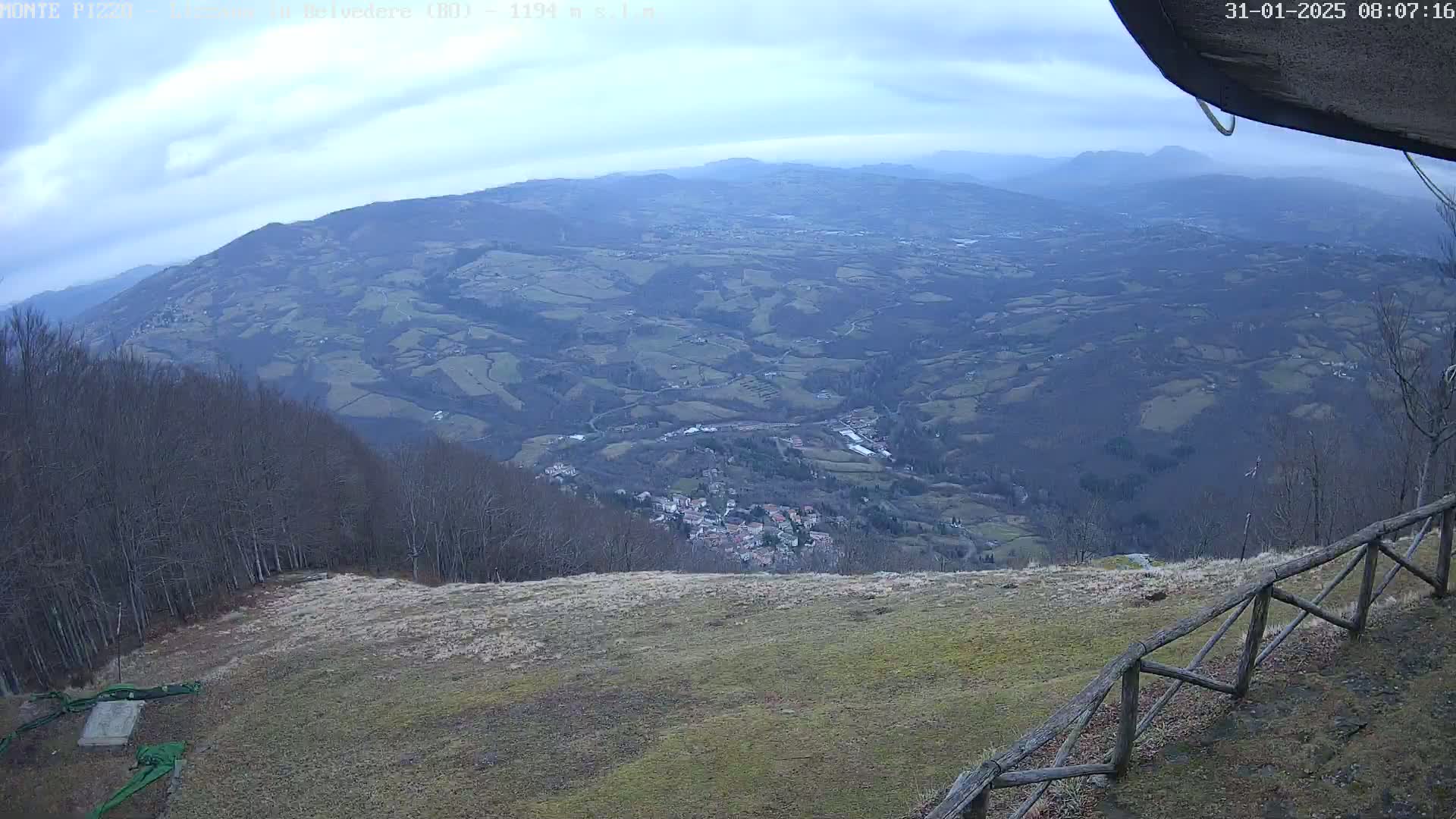 A high-angle view on an overcast day shows a vast, hilly landscape with a small village nestled in a valley below, viewed from a grassy area with a wooden fence.