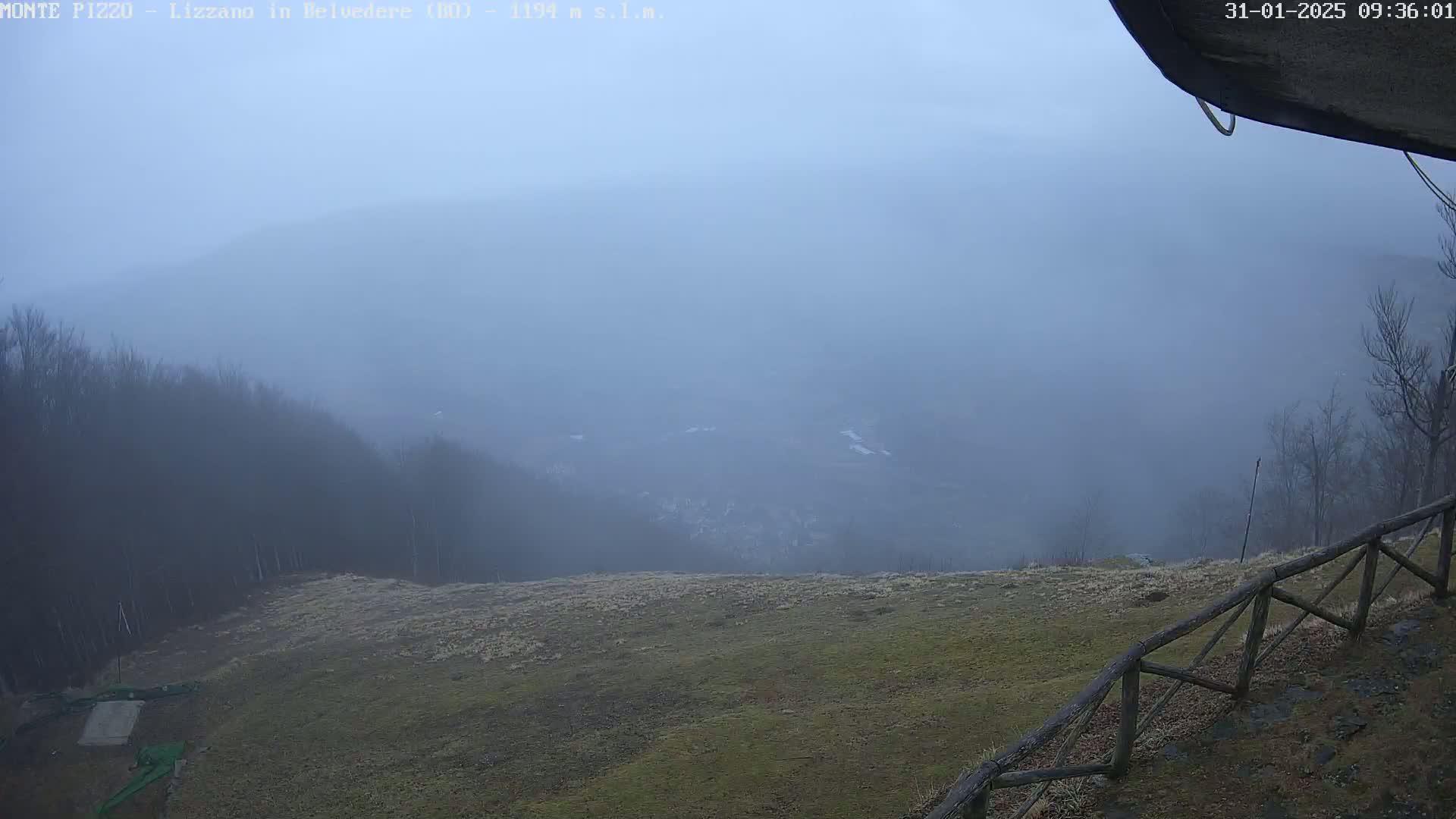 A foggy view from a high vantage point reveals a grassy hilltop with a wooden fence, overlooking a distant, obscured valley and tree-covered hills.