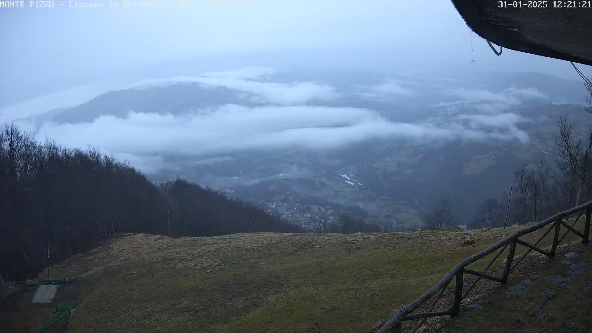 A foggy valley stretches below a mountaintop, where a wooden fence borders a grassy area.