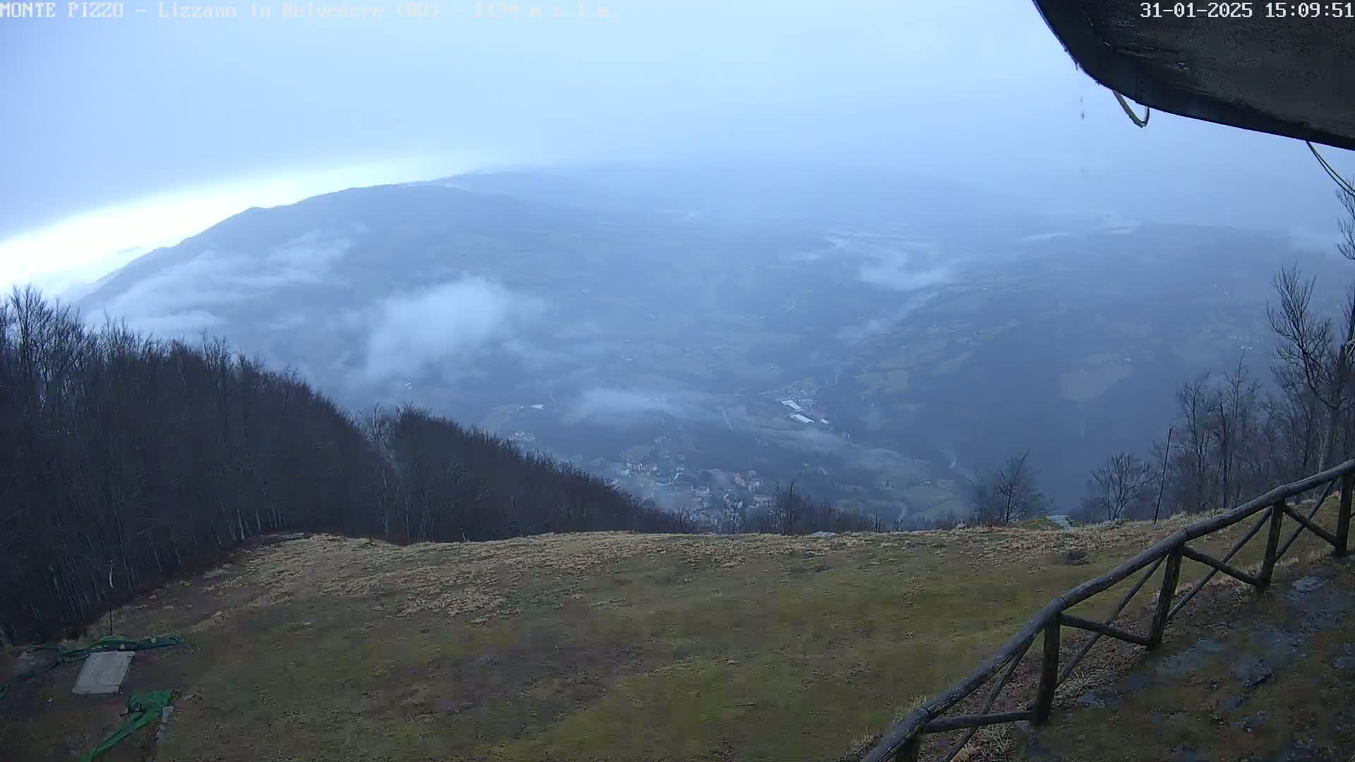 A high-angle view shows a misty valley below a mountaintop with bare trees and a wooden fence, under an overcast sky.
