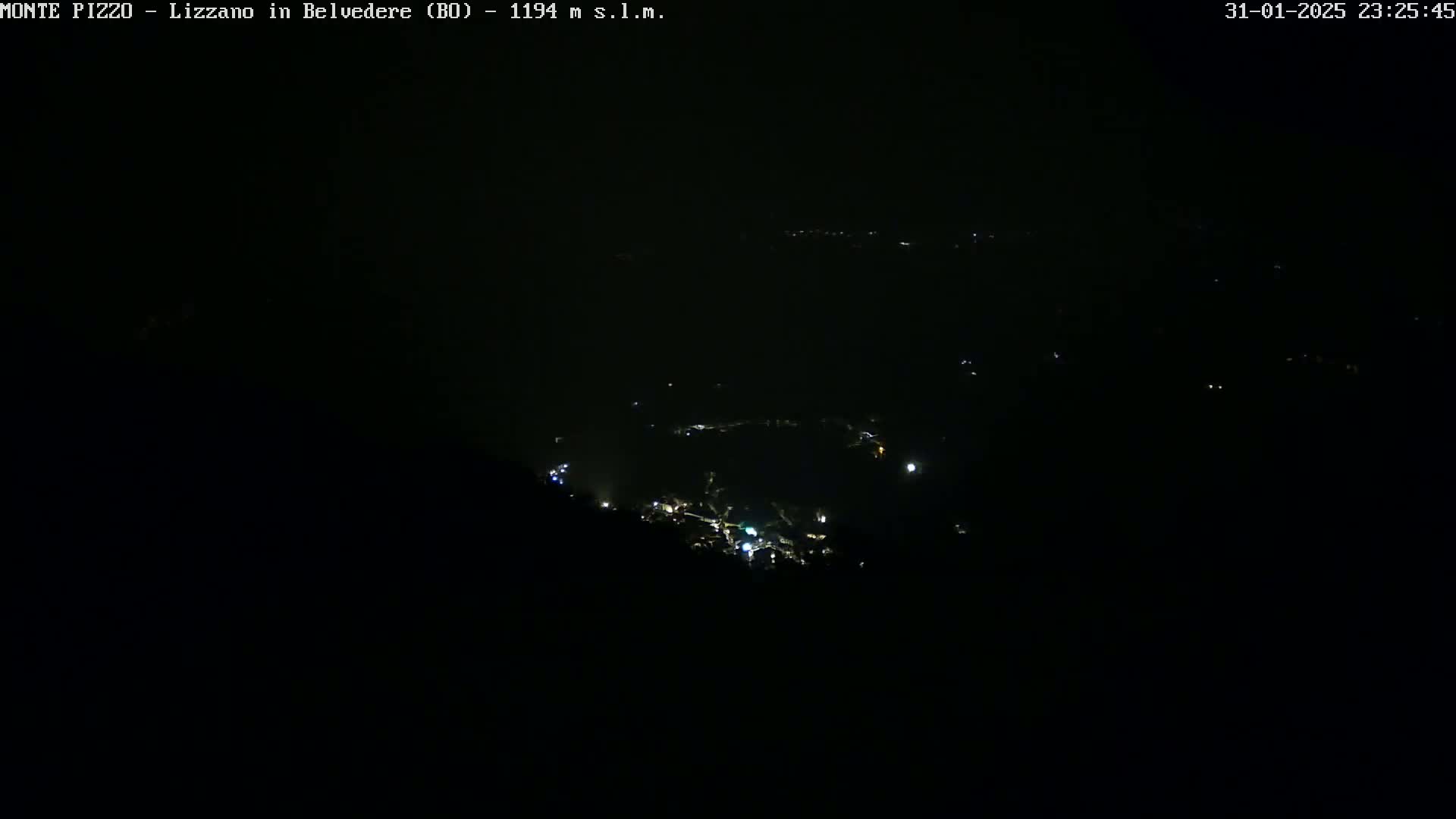 A nighttime, high-angle view shows a dark landscape with a few scattered lights from a distant town below.