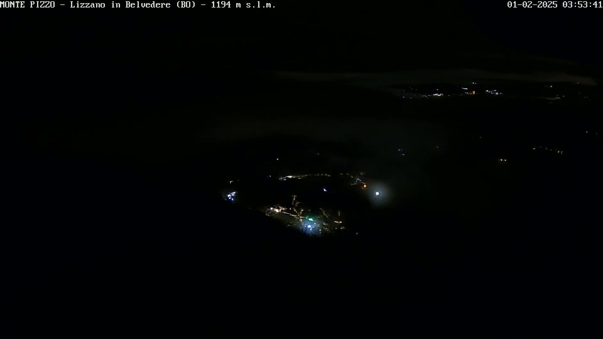 A nighttime, high-angle view shows a small town partially obscured by fog or low clouds, with scattered lights illuminating the scene.