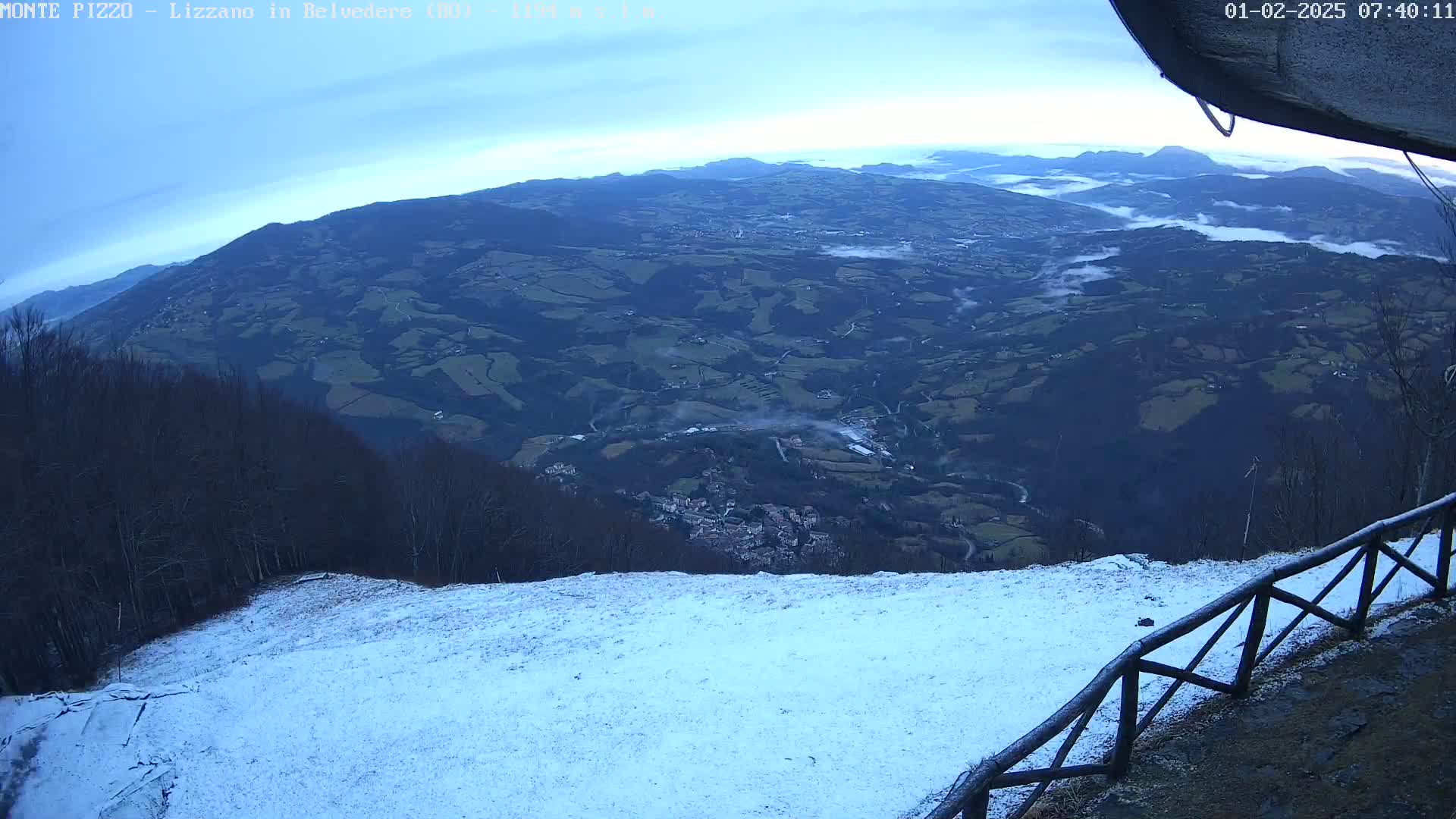 A snow-covered hillside overlooks a valley with low-lying fog under a mostly clear sky.