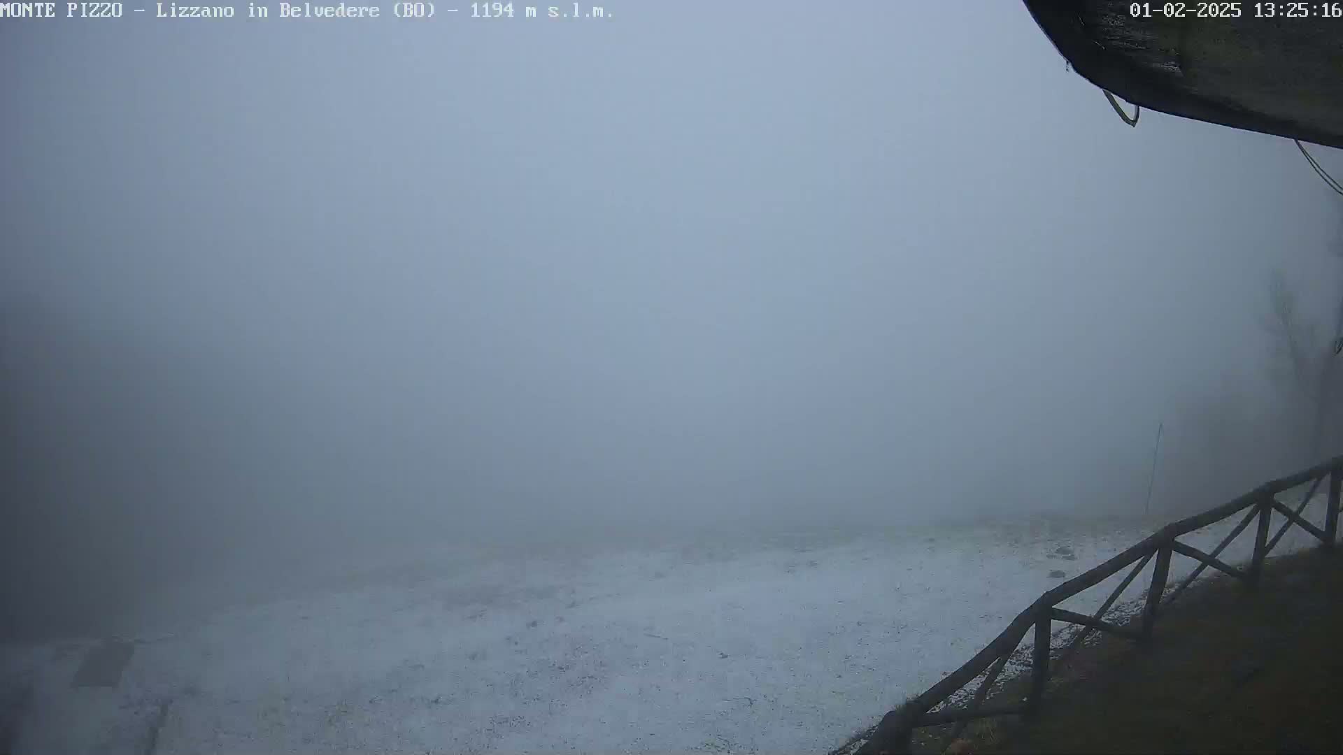 A snow-covered ground is visible through a thick fog from a high vantage point, with a wooden railing in the lower right corner.