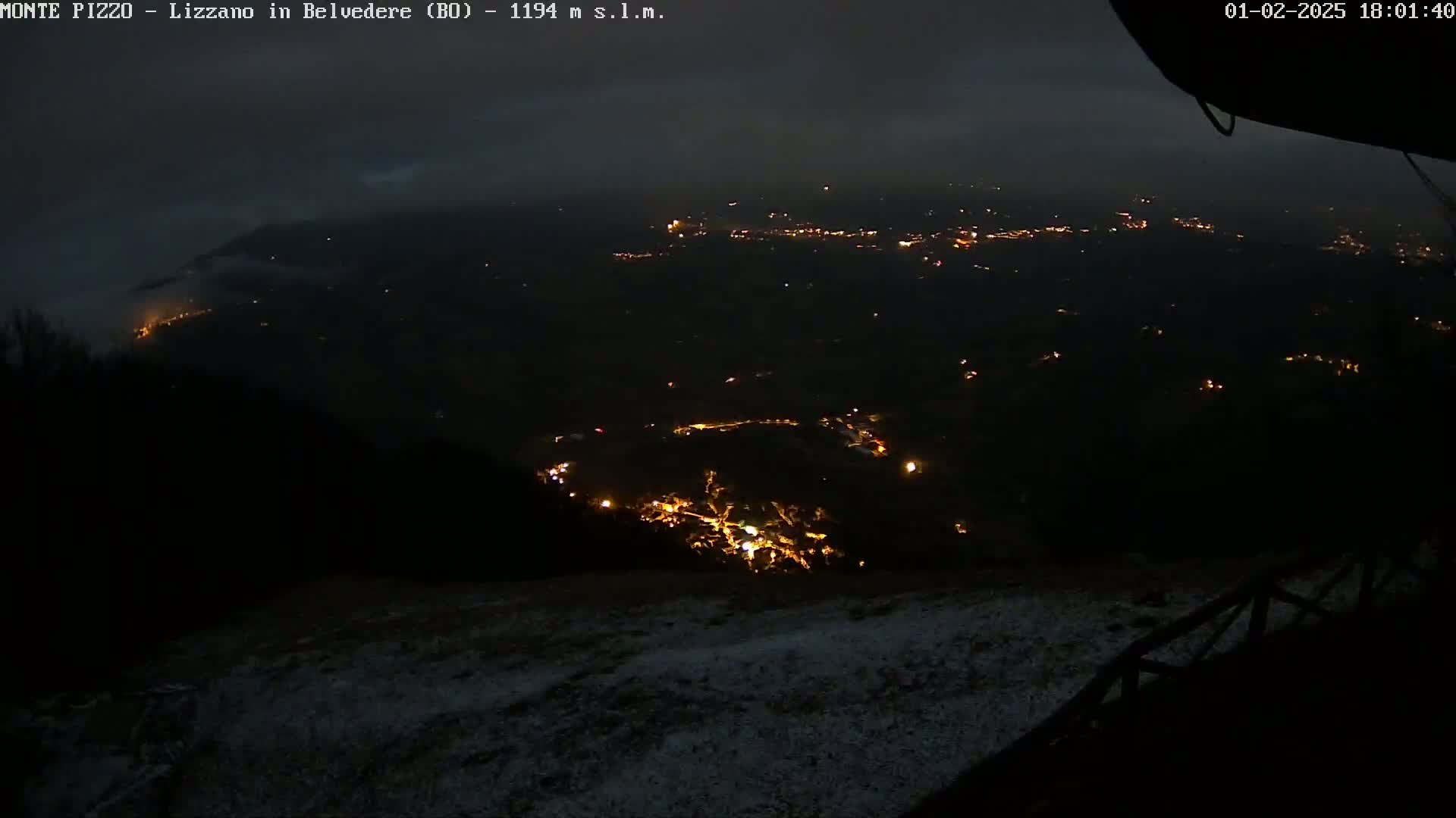 A nighttime, snowy landscape shows a valley below with scattered lights from a town or village under a cloudy sky.