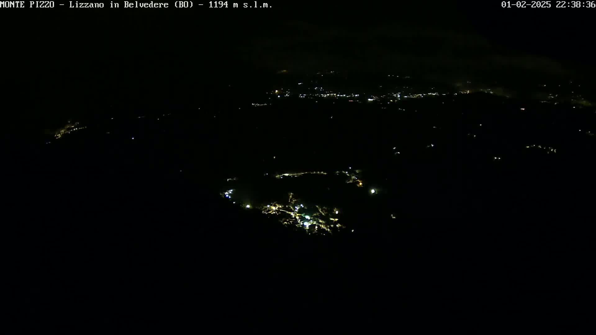 A nighttime, aerial view shows a dark landscape speckled with faint lights from a distant town under a cloudy sky.