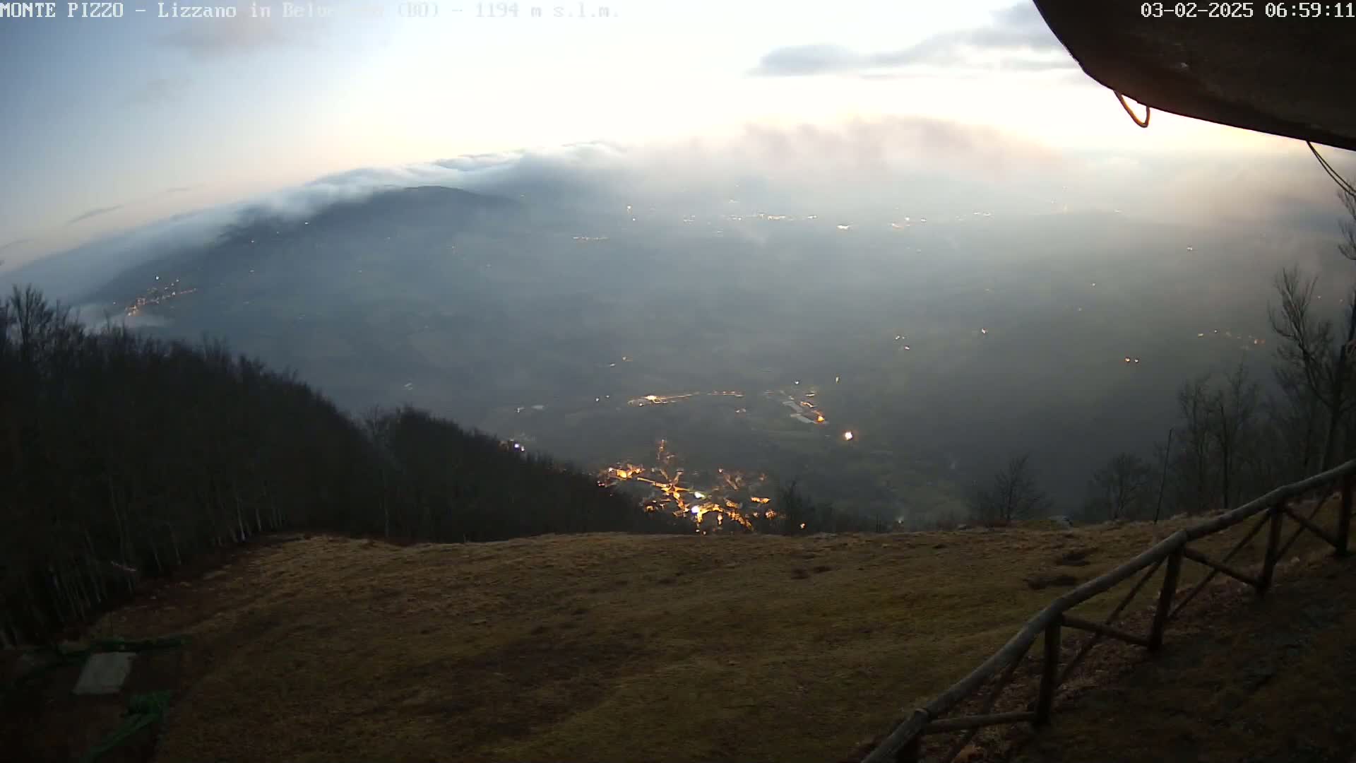 A high-angle, twilight view shows a valley filled with low clouds and scattered lights, seen from a hilltop with a wooden fence and bare trees.