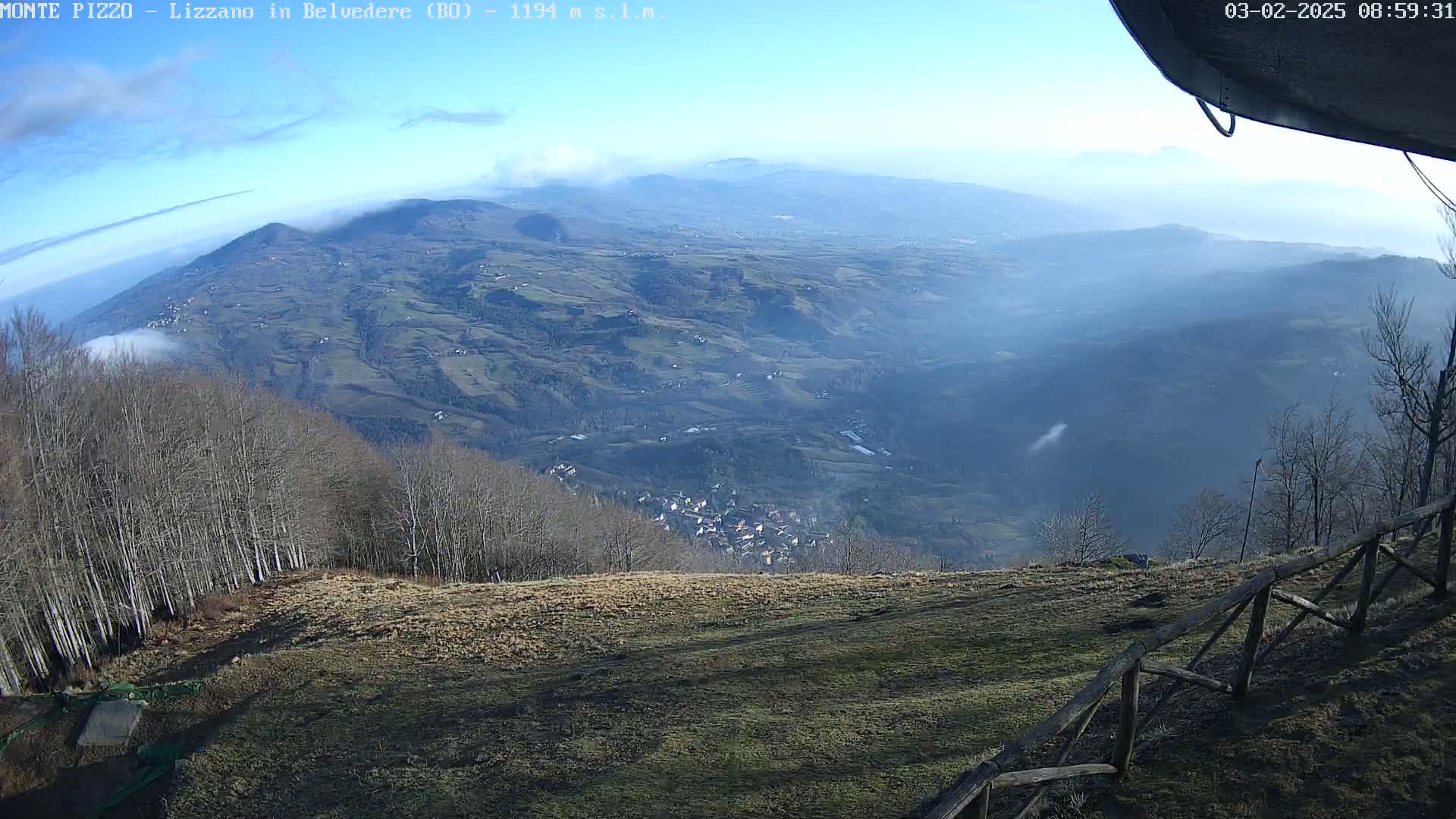 A hazy, sunny view from a mountaintop reveals a valley dotted with small villages and fields, bordered by leafless trees in the foreground.