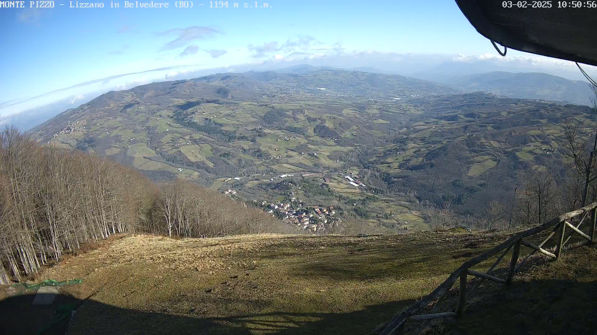 A high-angle view on a sunny day shows a vast, hilly landscape with a small town nestled in the valley below, surrounded by bare winter trees and farmland, extending to a layer of clouds in the distance.