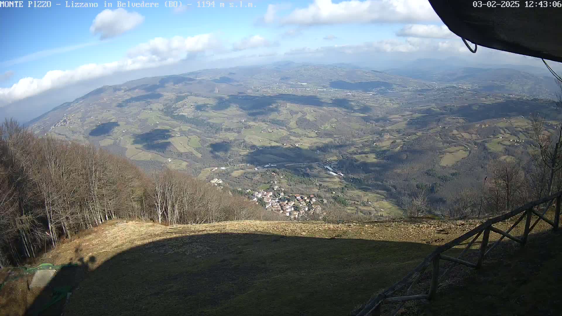A partly sunny day reveals a panoramic view of a valley with a small village nestled among rolling hills and sparse, leafless trees, seen from a high vantage point.