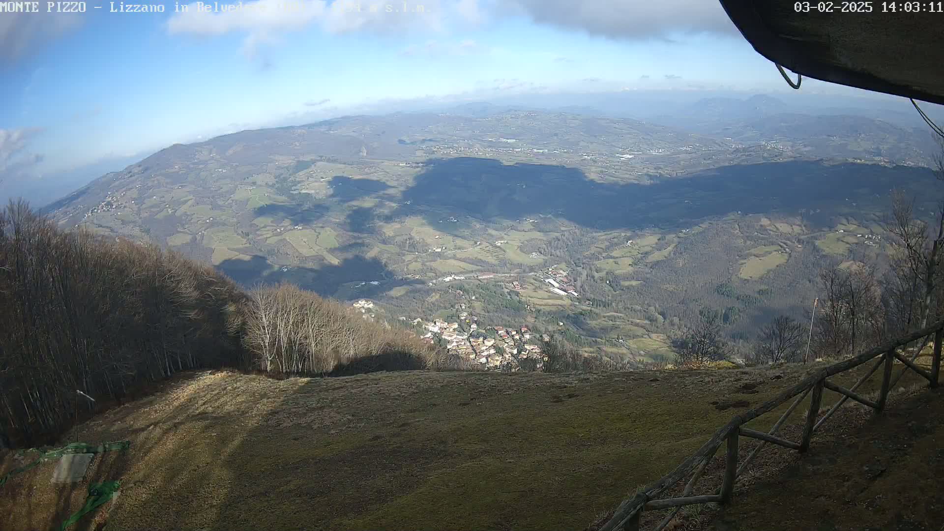 A high-angle, sunny view of a vast valley dotted with small villages, partially obscured by the shadows of clouds, with a foreground of a grassy slope and wooden fence.