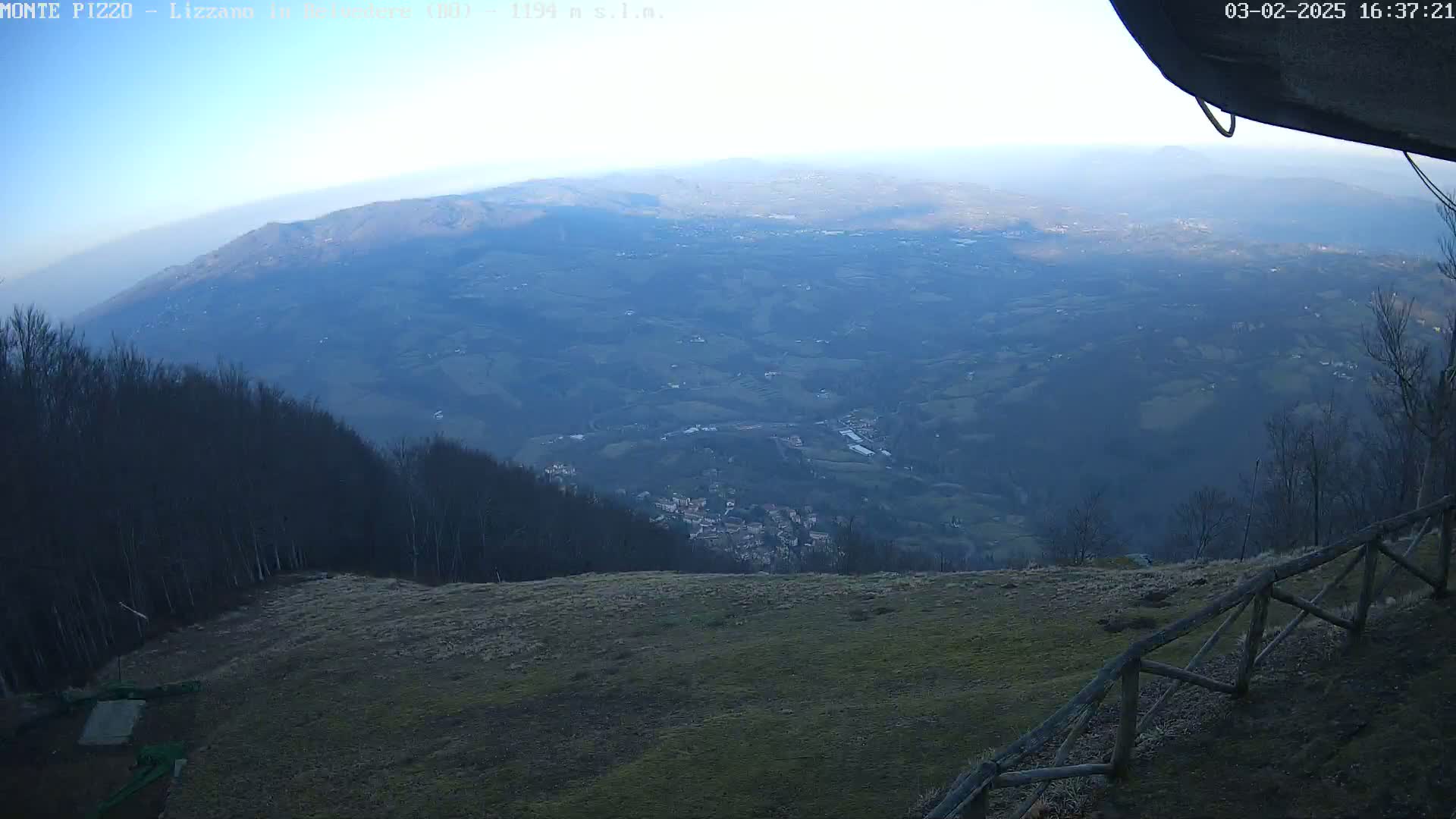A hazy, distant view of a valley and town from a mountaintop, with a wooden fence in the foreground under a clear sky.