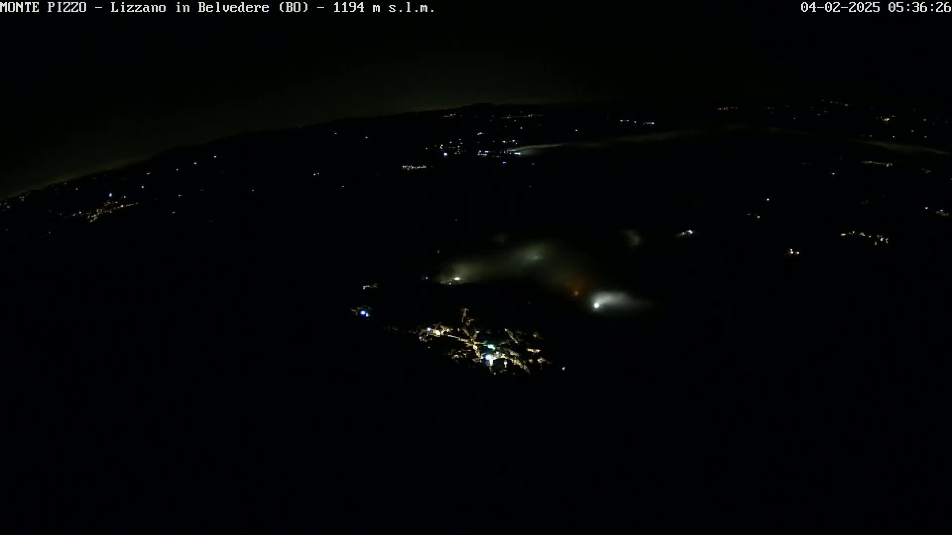 A nighttime, high-angle view of a town partially obscured by fog or low clouds, with scattered lights visible below.
