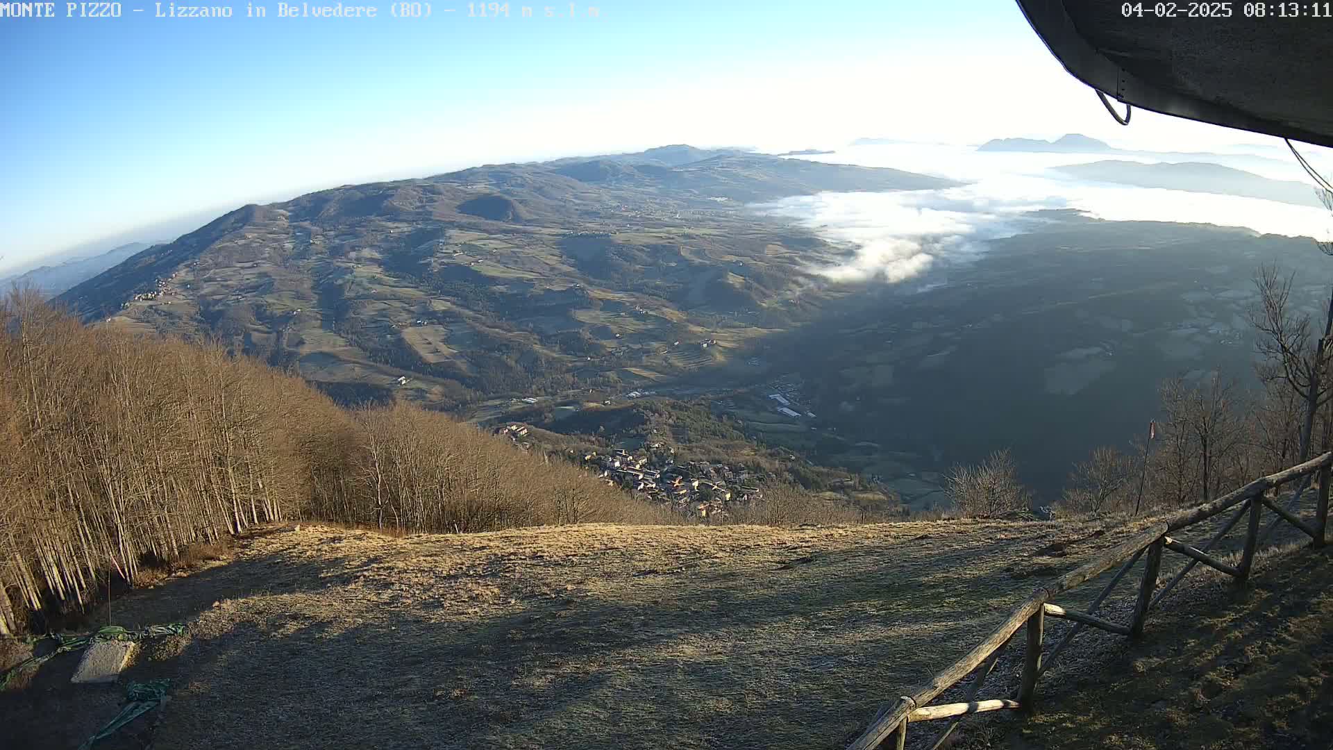 A high-altitude view of a valley blanketed in low-lying fog under a clear blue sky, showing a small village nestled amongst rolling hills and leafless trees.