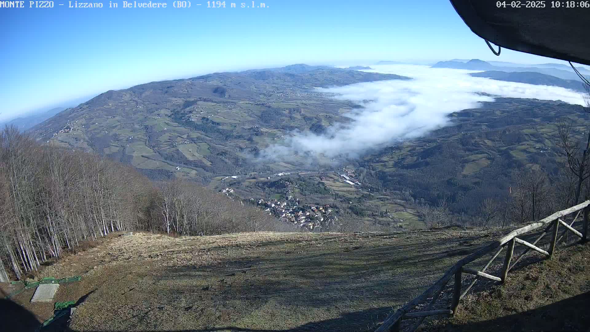 A high-angle, long shot on a clear, sunny day shows a valley filled with low-lying clouds, a small town nestled among fields and forests, and a wooden fence in the foreground.