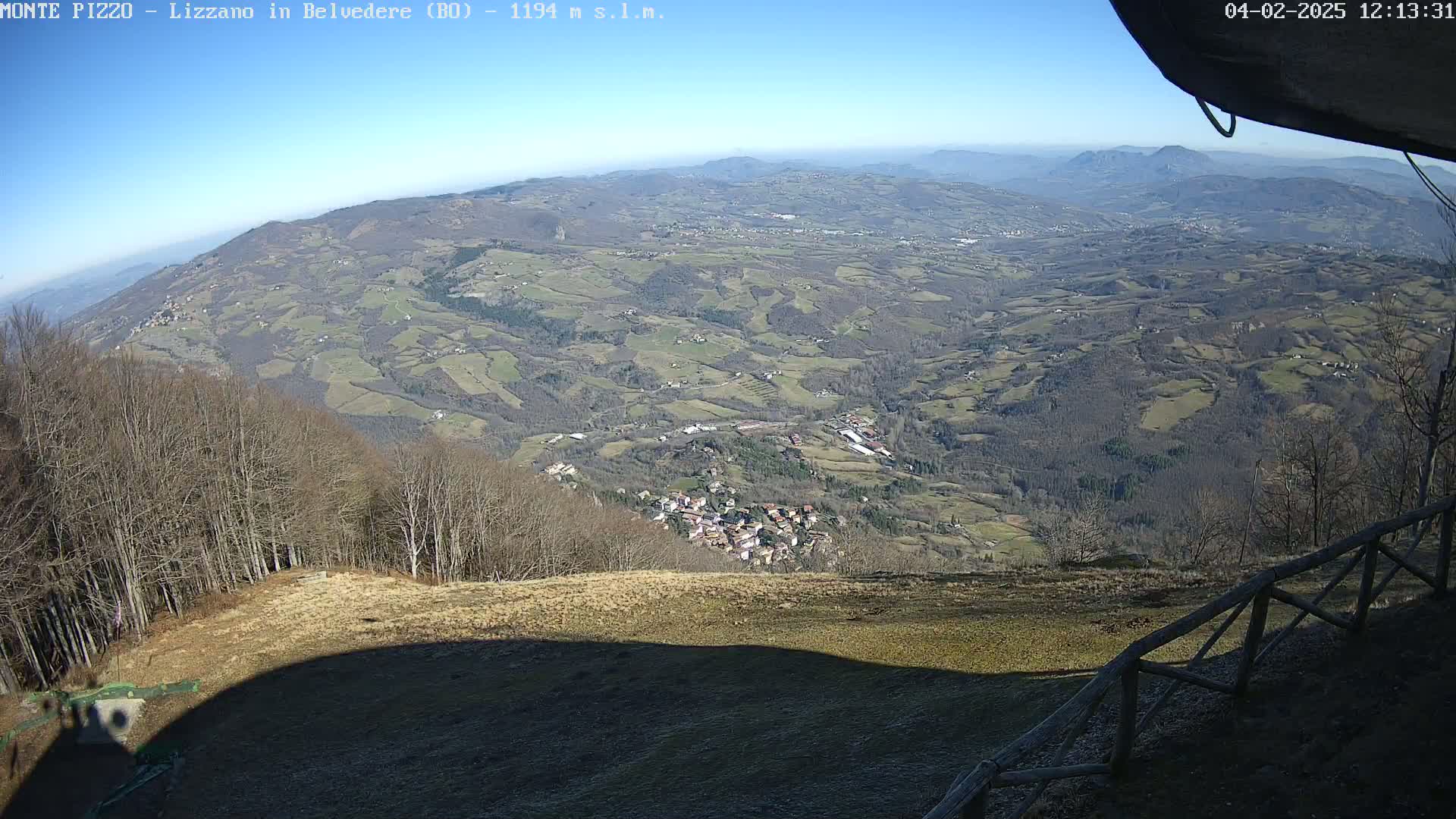 A high-angle, long shot on a sunny day shows a vast expanse of farmland and a small village nestled in a valley, viewed from a wooded mountaintop with a wooden fence in the foreground.