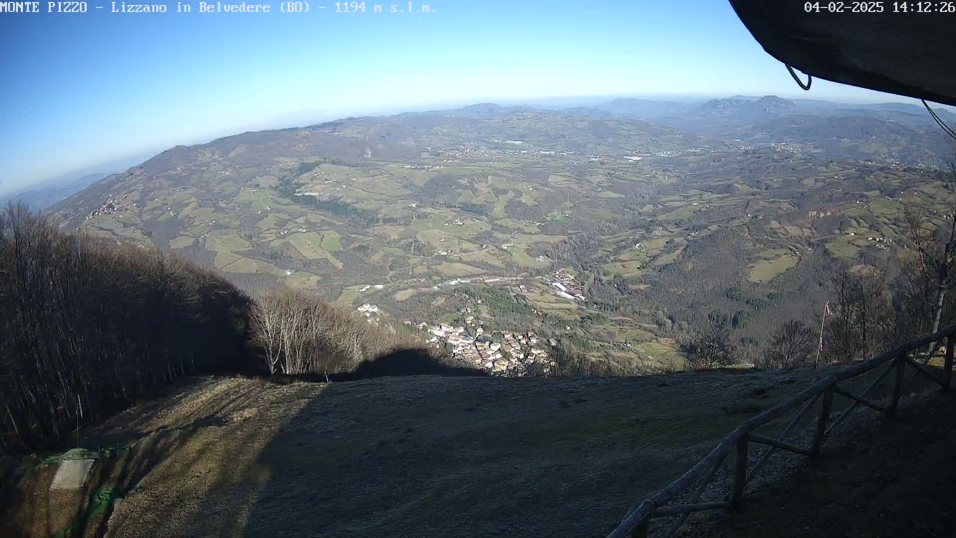 A high-angle, long shot on a sunny day shows a sprawling valley with a small town nestled within, surrounded by rolling hills and forests, viewed from a mountaintop vantage point.