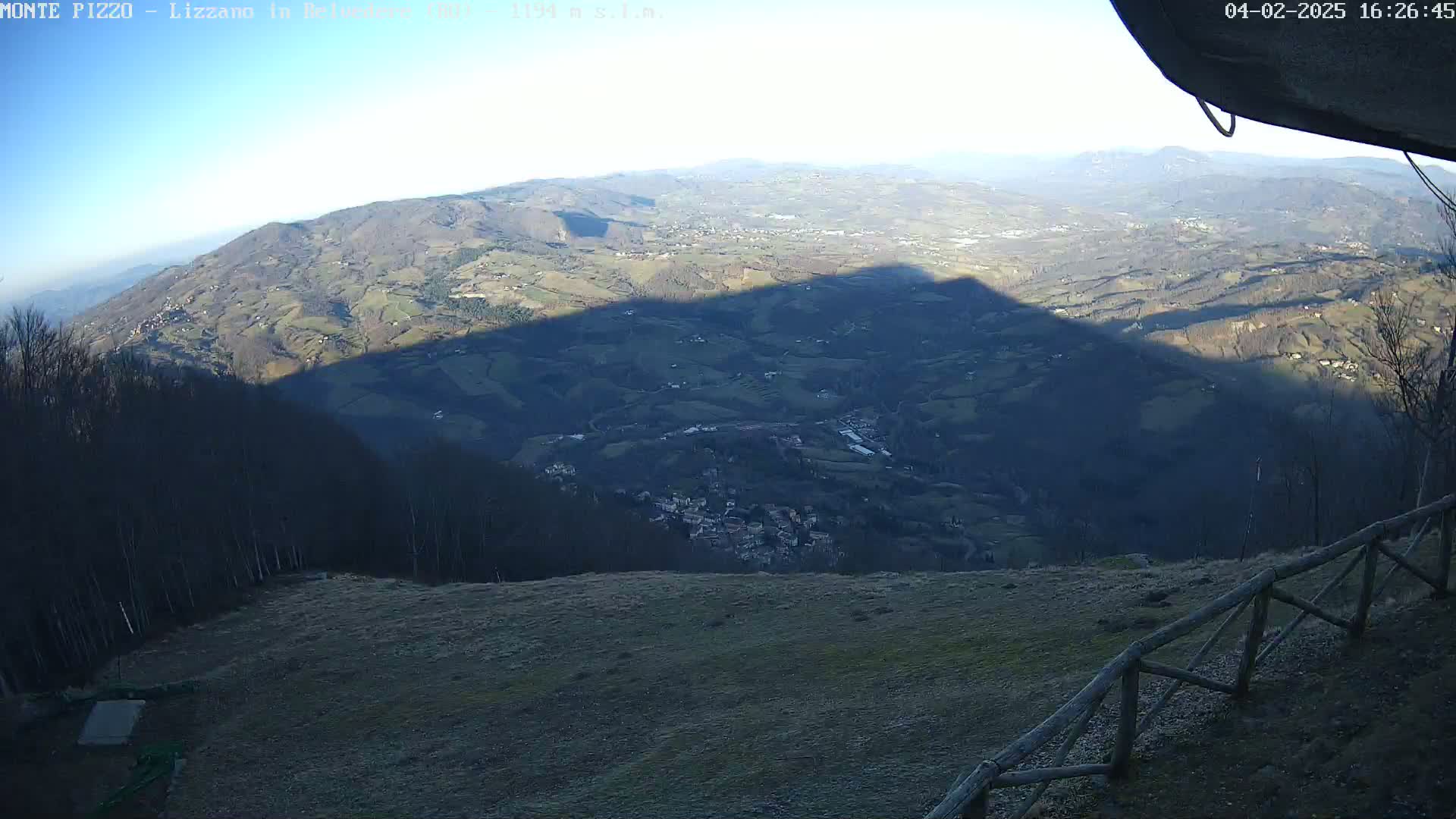 A panoramic view from a high vantage point shows a vast valley with a small town nestled within, partially obscured by the long shadow of a mountain under a clear, bright sky.