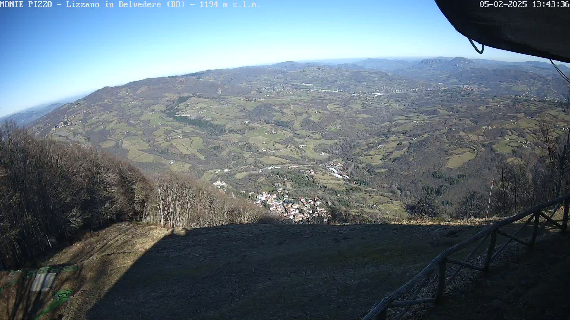 A high-angle, long shot on a sunny day shows a vast, hilly landscape dotted with small towns and villages, partially obscured by a foreground of bare trees and a wooden fence.