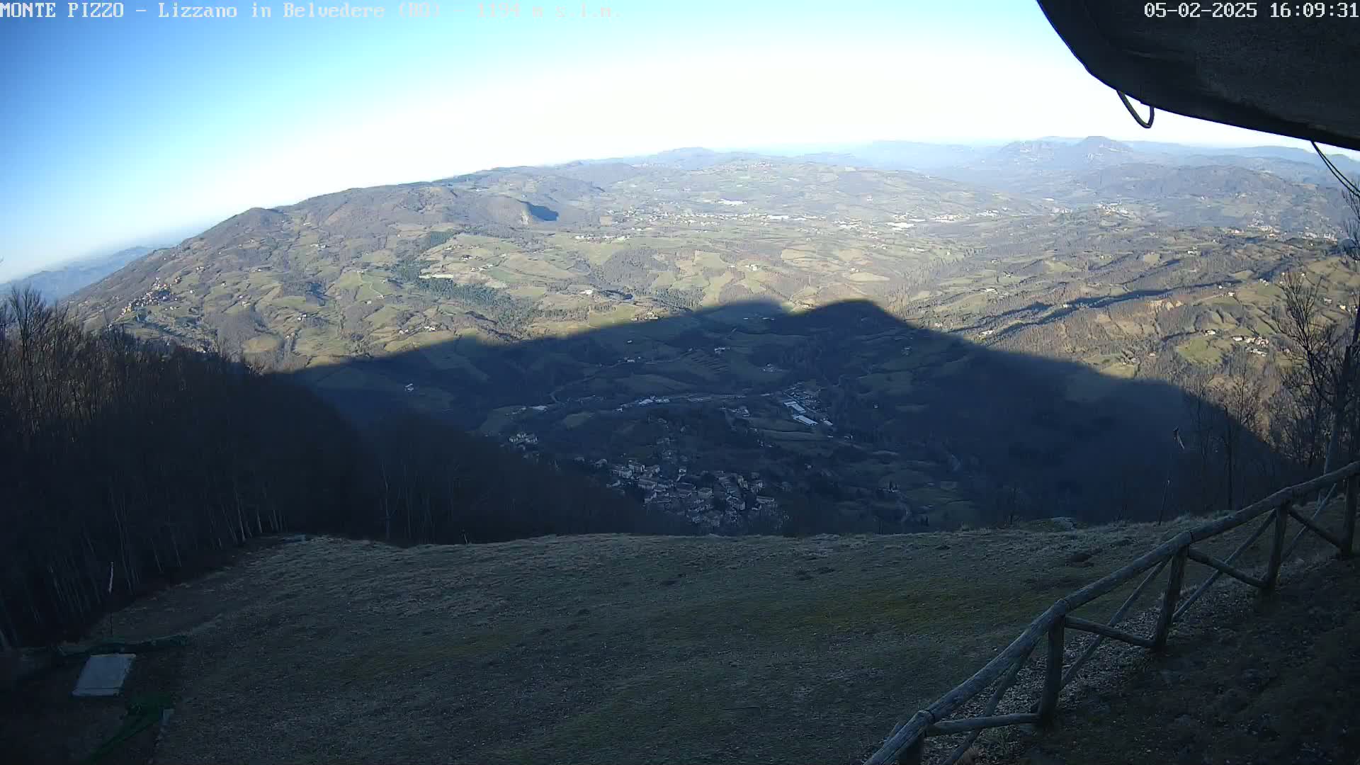 A high-angle, long shot shows a sprawling, hilly landscape under a clear, sunny sky, with a large shadow cast across the valley below.