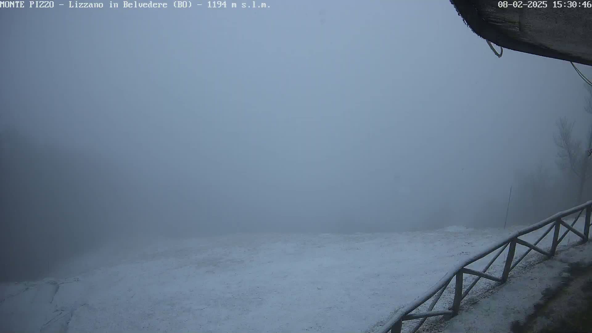 A snow-covered landscape is shrouded in thick fog, with a wooden fence visible in the foreground.