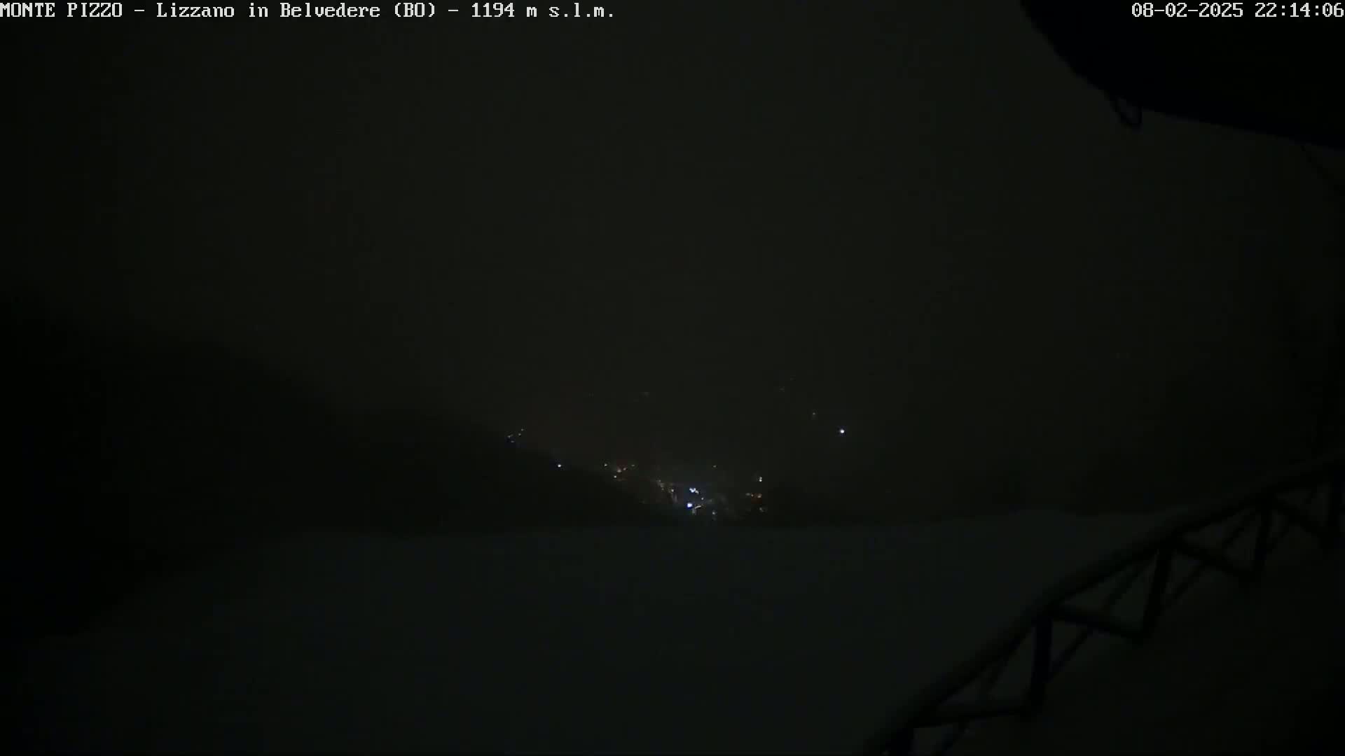 A dark, snowy nighttime scene shows a few distant lights in a valley below.