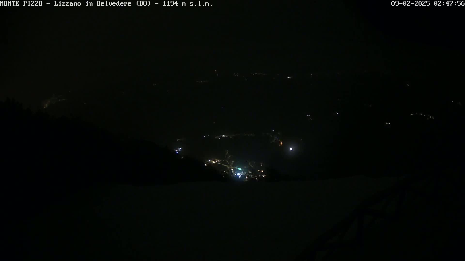 A nighttime, high-angle view shows a valley with sparsely lit buildings below a dark sky.