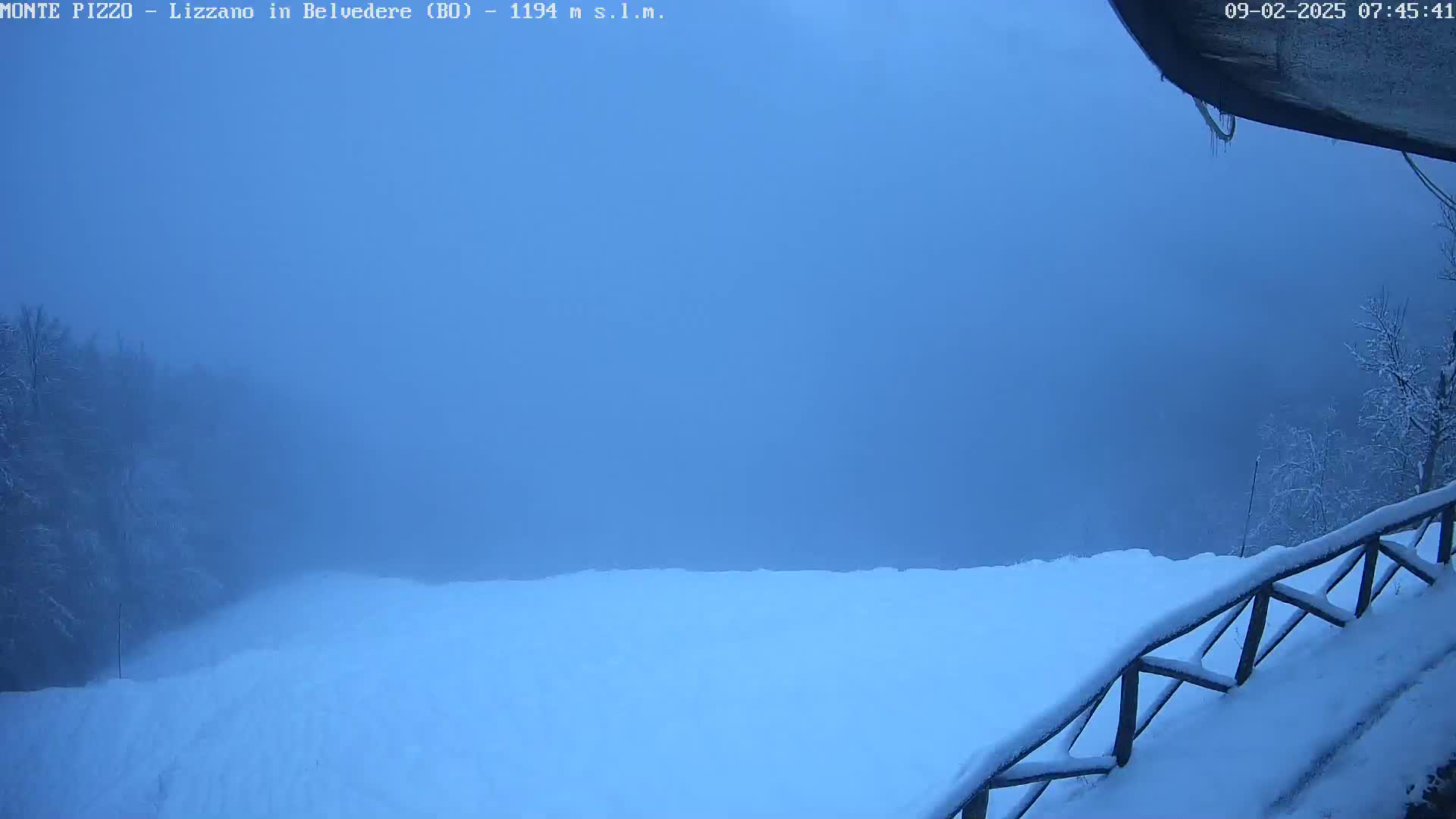 A snow-covered landscape is shrouded in dense fog, with a wooden fence partially visible in the lower right corner.