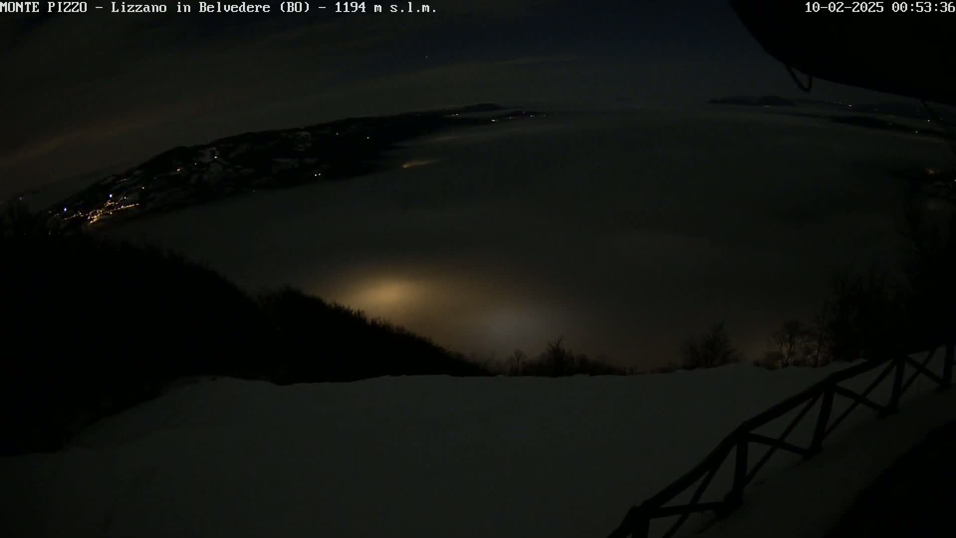 A nighttime view from a snowy, high vantage point shows a valley filled with fog, with distant lights faintly visible below.