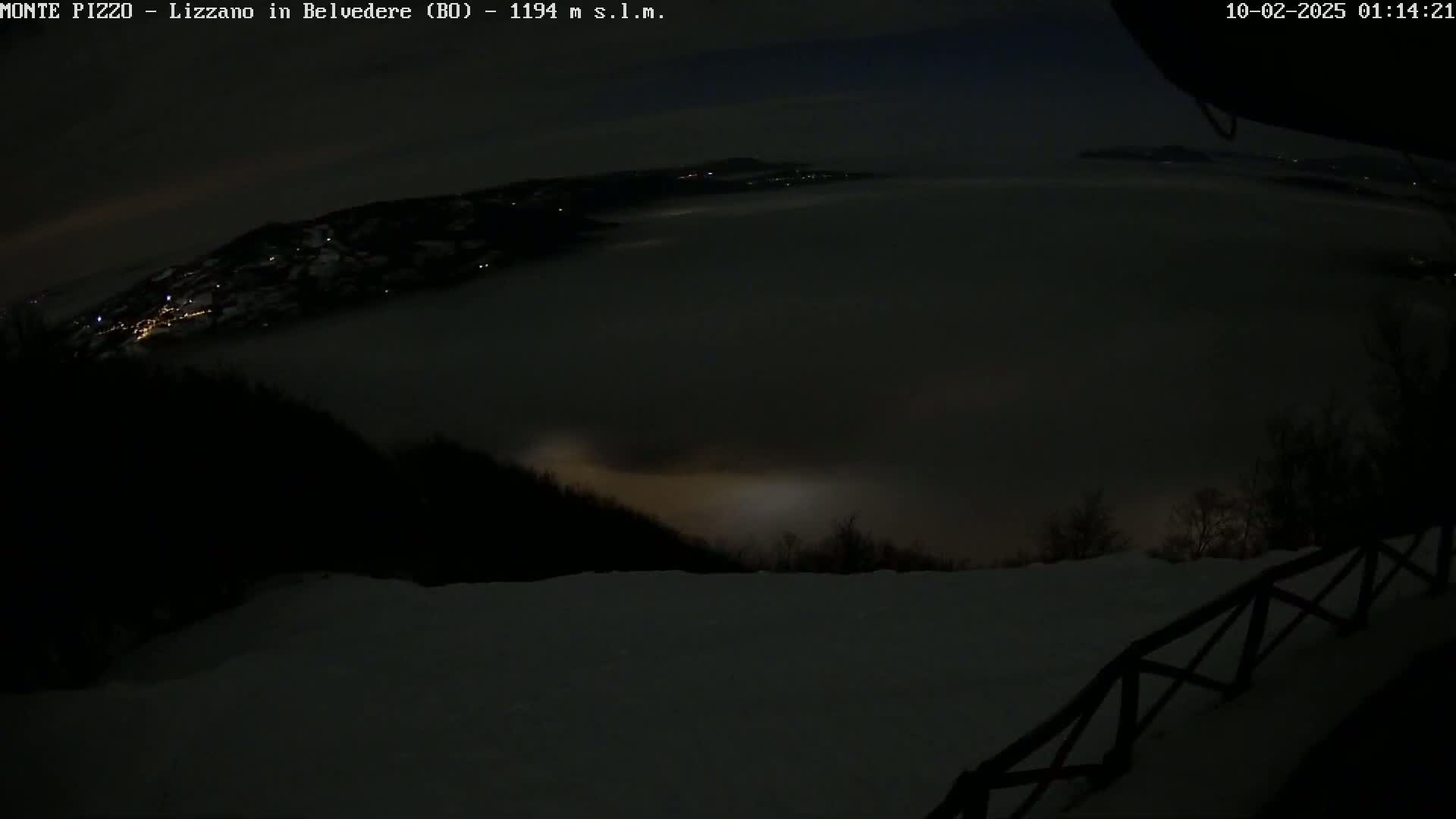 A nighttime view from a snowy, high vantage point shows a valley covered in fog with dimly lit settlements visible in the distance under a dark sky.