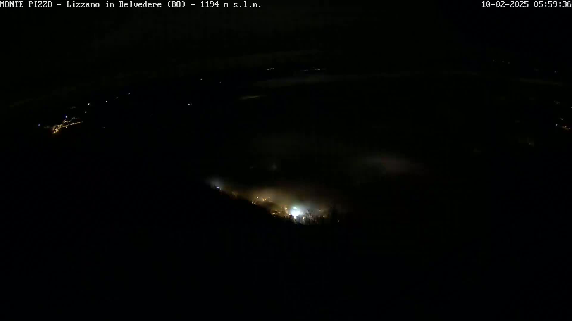 A nighttime, high-angle view shows a low-lying layer of fog or clouds obscuring a dimly lit town below.