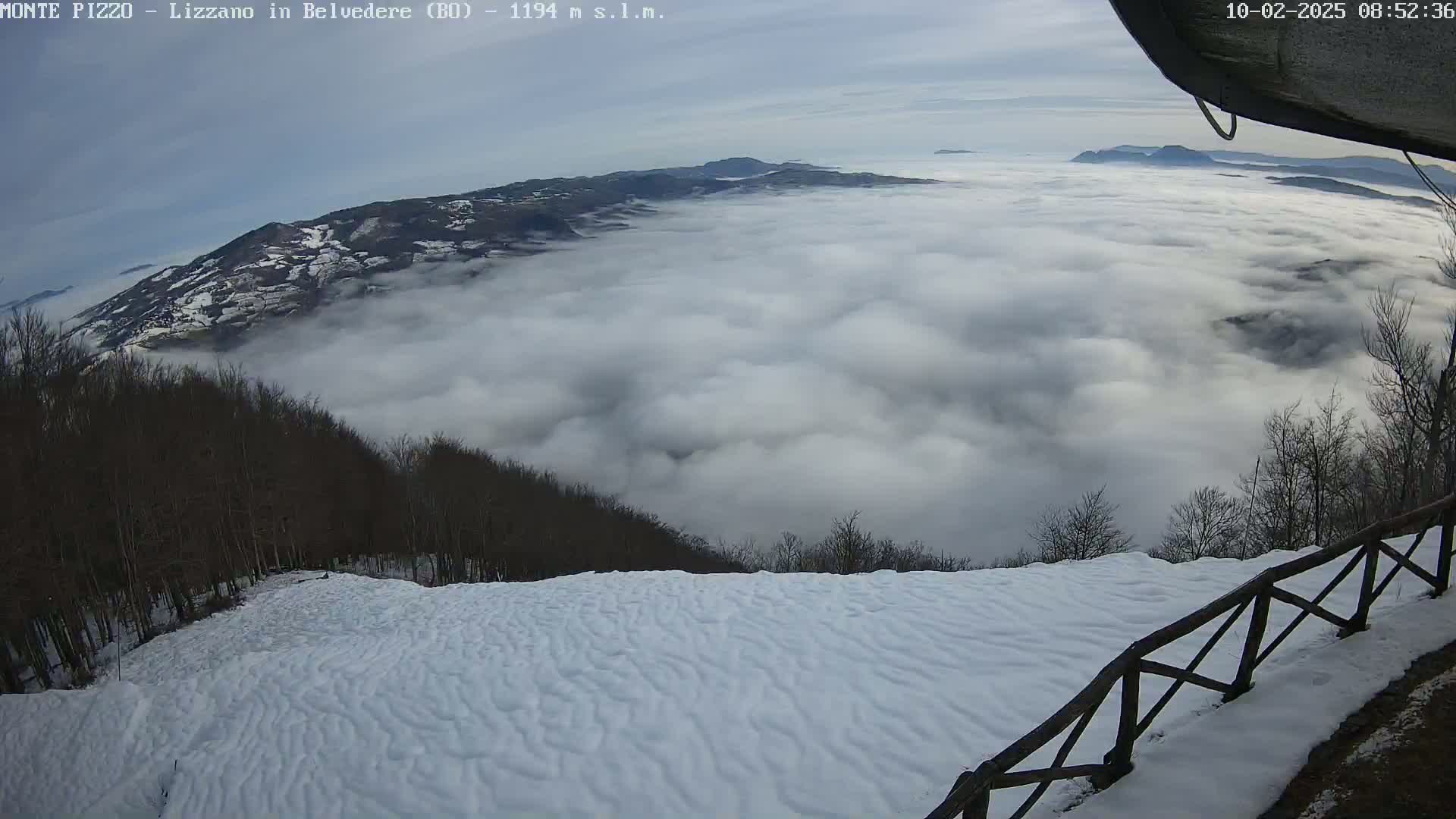 A snowy landscape overlooks a vast expanse of clouds below, with bare trees lining the hillside.