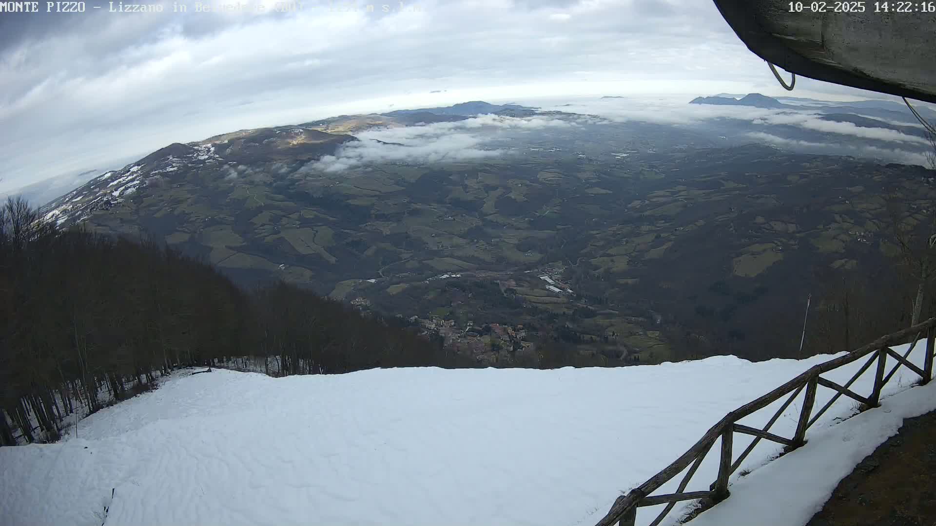 A snowy mountaintop view reveals a valley covered in a low layer of clouds, with distant mountains visible beyond.