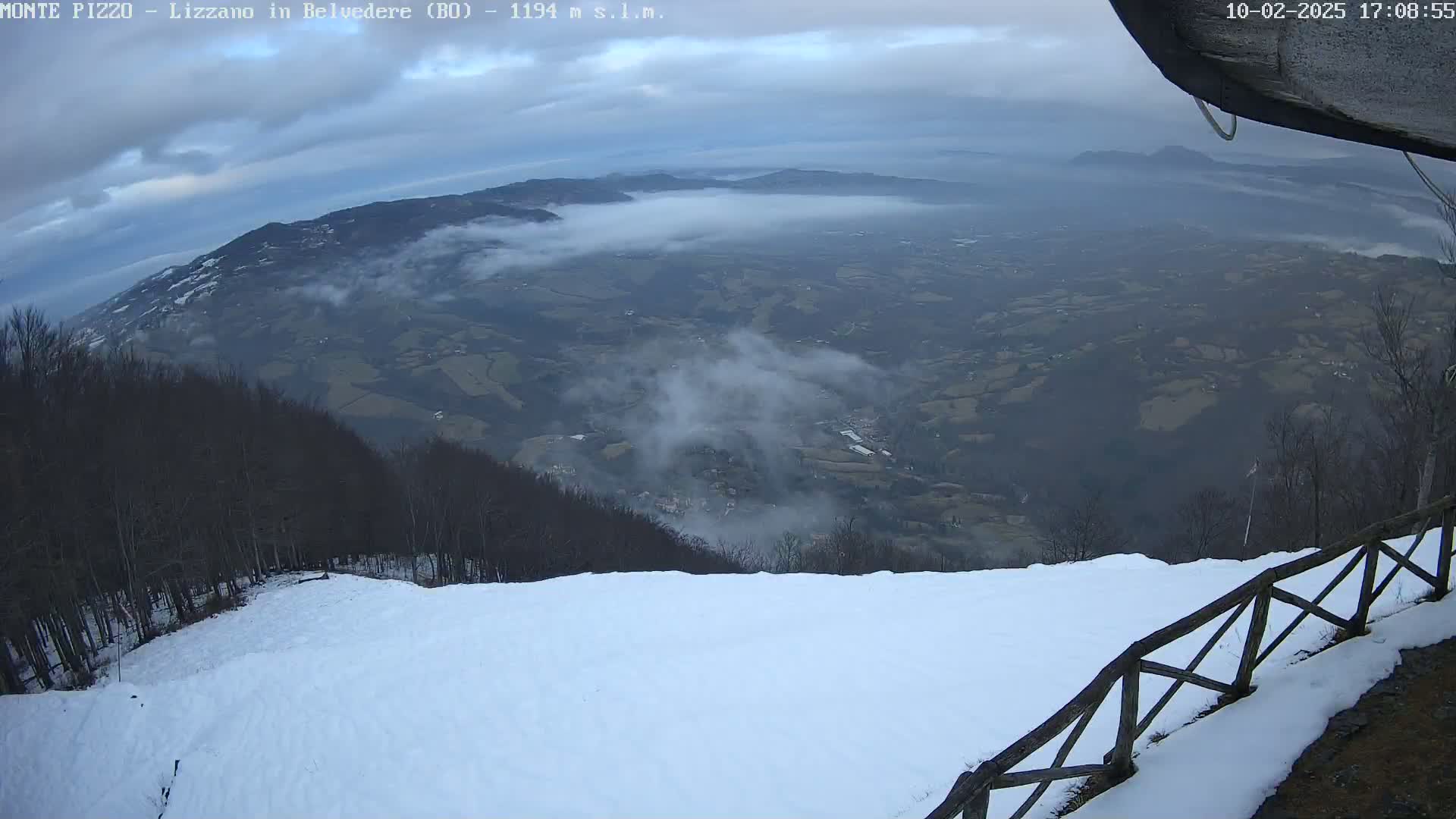 A snow-covered ski slope overlooks a valley shrouded in fog under a cloudy sky.
