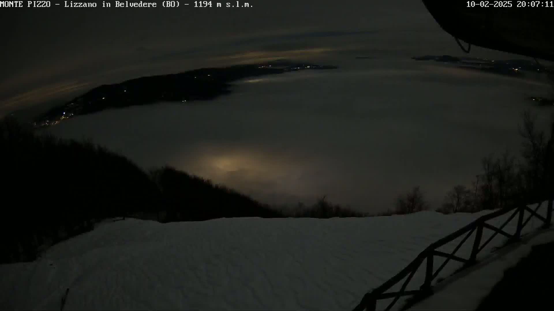 A nighttime view from a snowy, elevated vantage point shows a low-lying fog covering a valley with distant lights visible through the fog.
