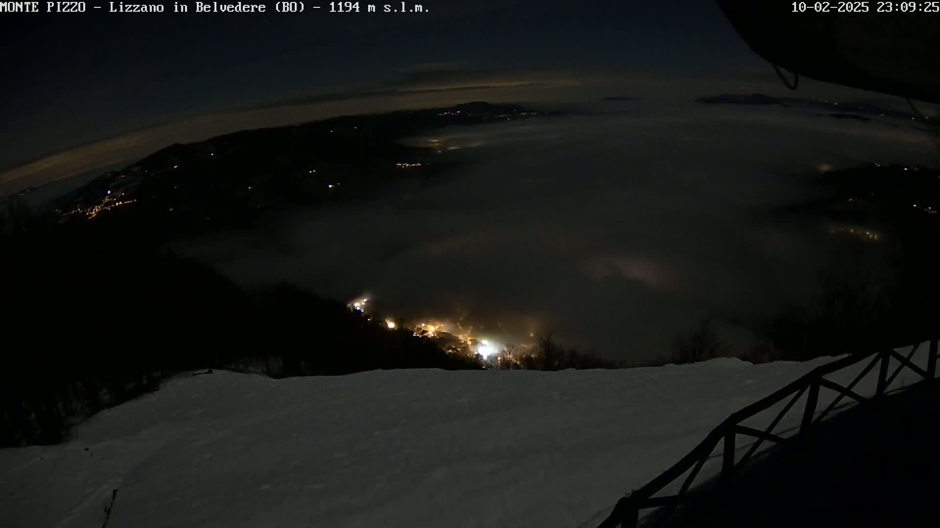 A nighttime, snowy landscape is viewed from a high vantage point, with a thick layer of fog or low clouds obscuring the lower areas and only some distant lights visible below.